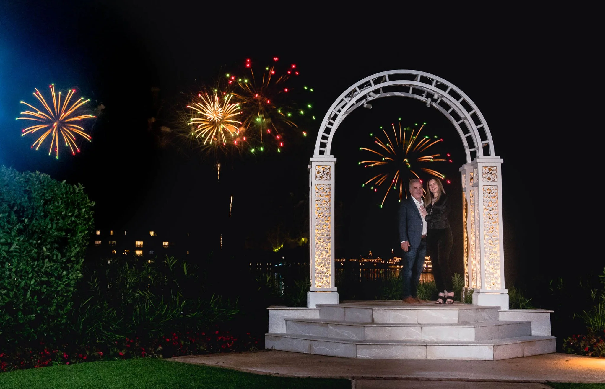 A couple standing under a lit white decorative arch at Disney's Picture Point at night with Magic Kingdom fireworks in the sky behind them.