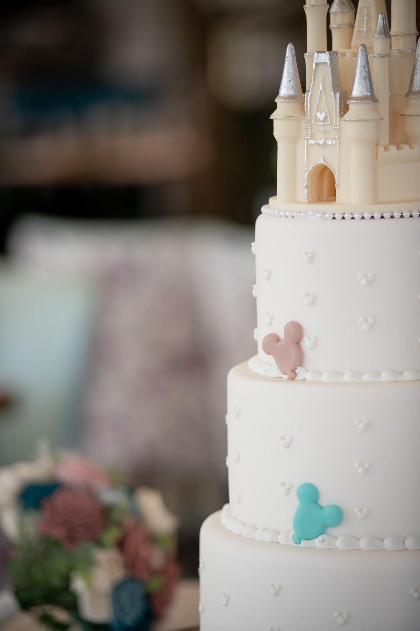 A multi-tiered wedding cake decorated with small Mickey Mouse head shapes and a pink and blue Mickey Mouse head on each tier, topped with a miniature castle. The cake has white icing, pearl-like border decorations, and silver accents on the castle.