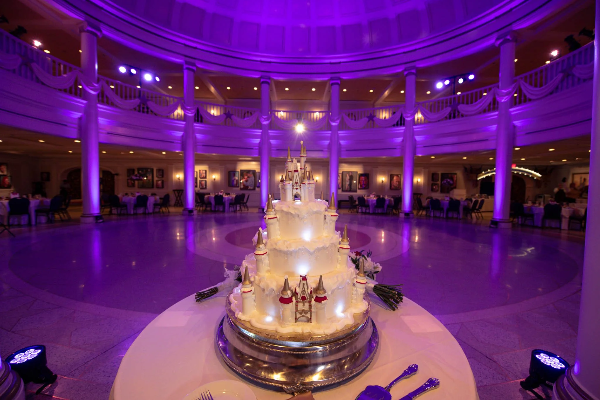 A multi-tiered cake with castle design decorations displayed on a table in the center of an elegant, purple-lit banquet hall at Epcot's American Adventure.