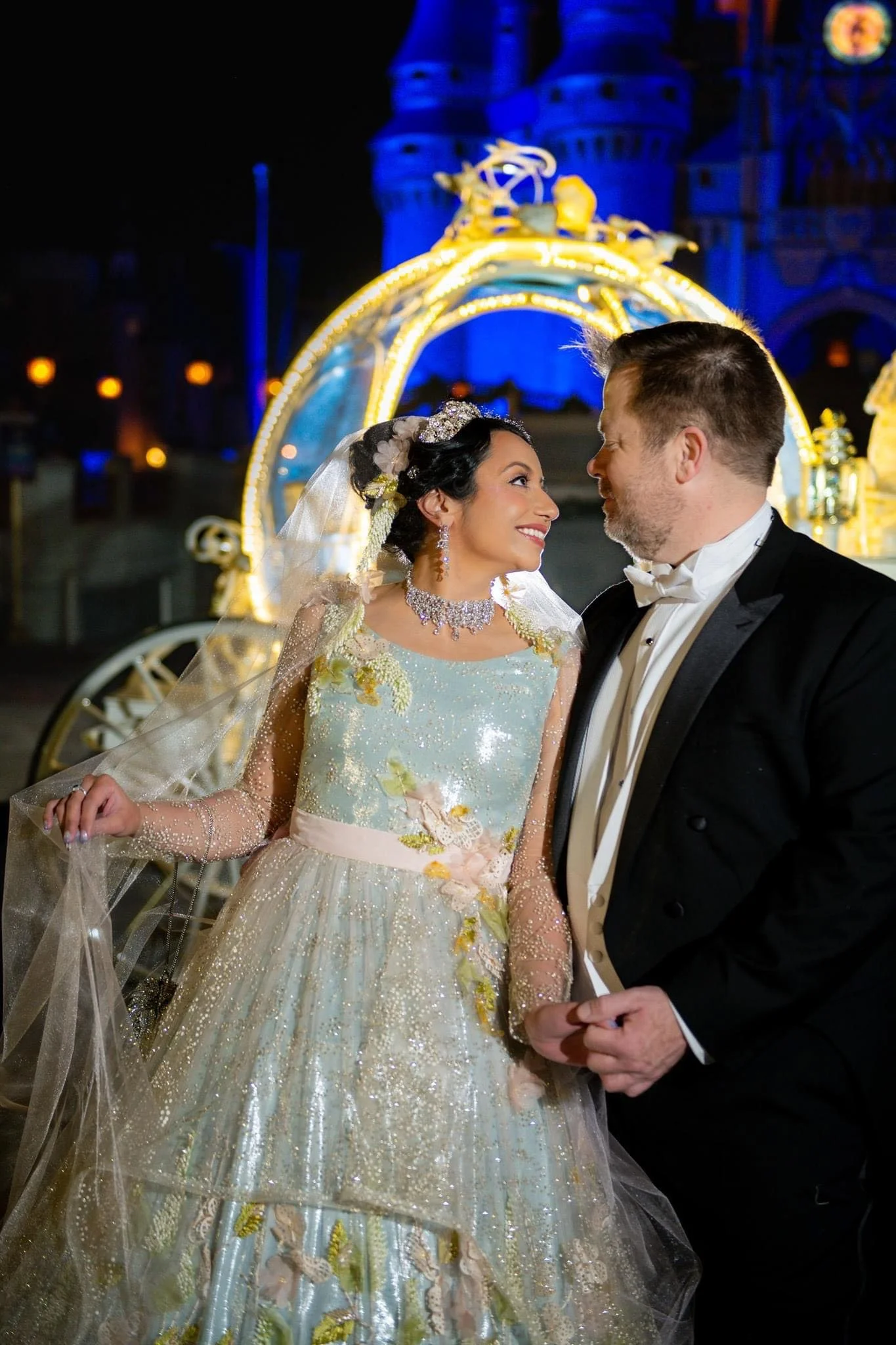 A newlywed couple in wedding attire, smiling at each other, standing in front of a decorated Magic Kingdom castle at night.