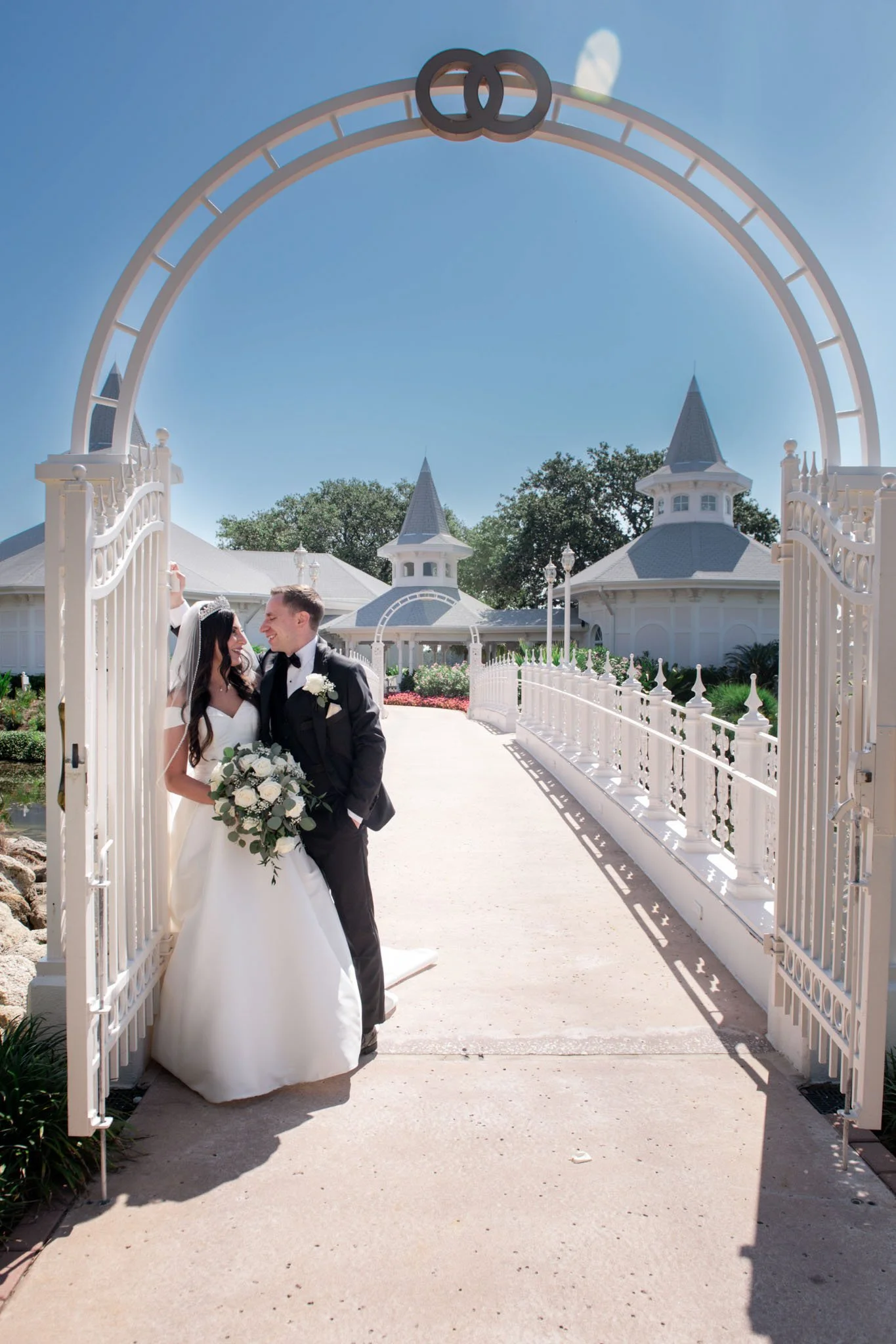 A bride and groom standing under an ornate gate at a wedding venue on a sunny day, surrounded by white buildings and lush greenery.