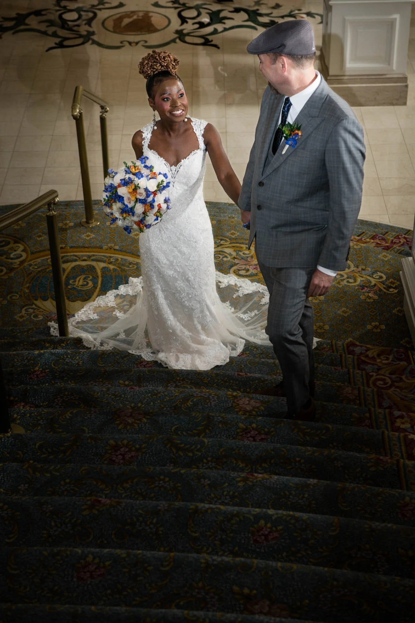 A bride in a white lace wedding gown holding a colorful bouquet, smiling and holding hands with a man in a gray plaid suit and gray flat cap on a staircase inside a building.
