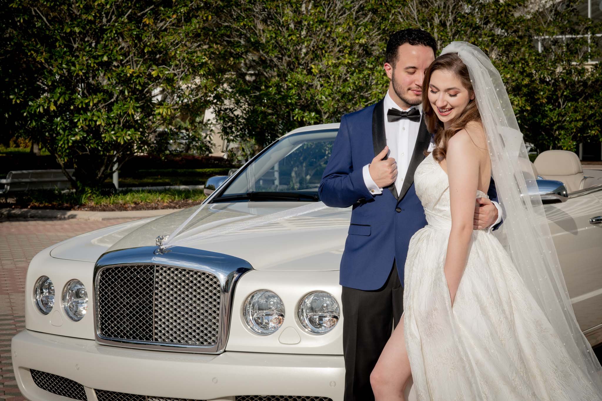 A bride and groom in wedding attire standing next to a white luxury car with ribbons, smiling and looking at each other.