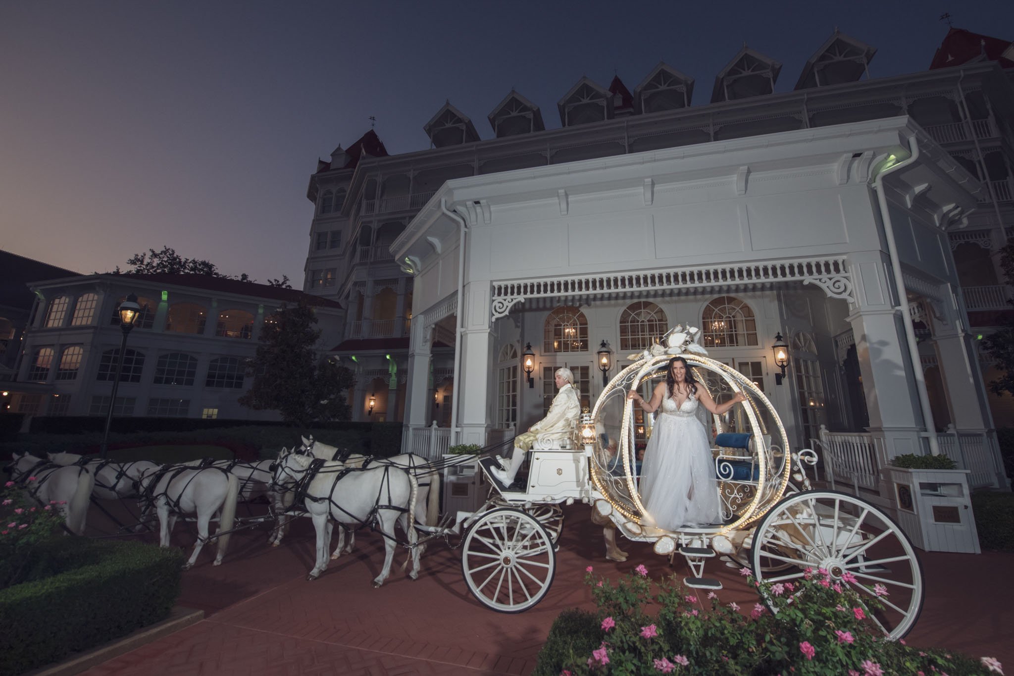 A bride in a white wedding dress sitting inside Cinderella's carriage lit with string lights, pulled by white horses, in front of a large Victorian-style hotel at dusk.