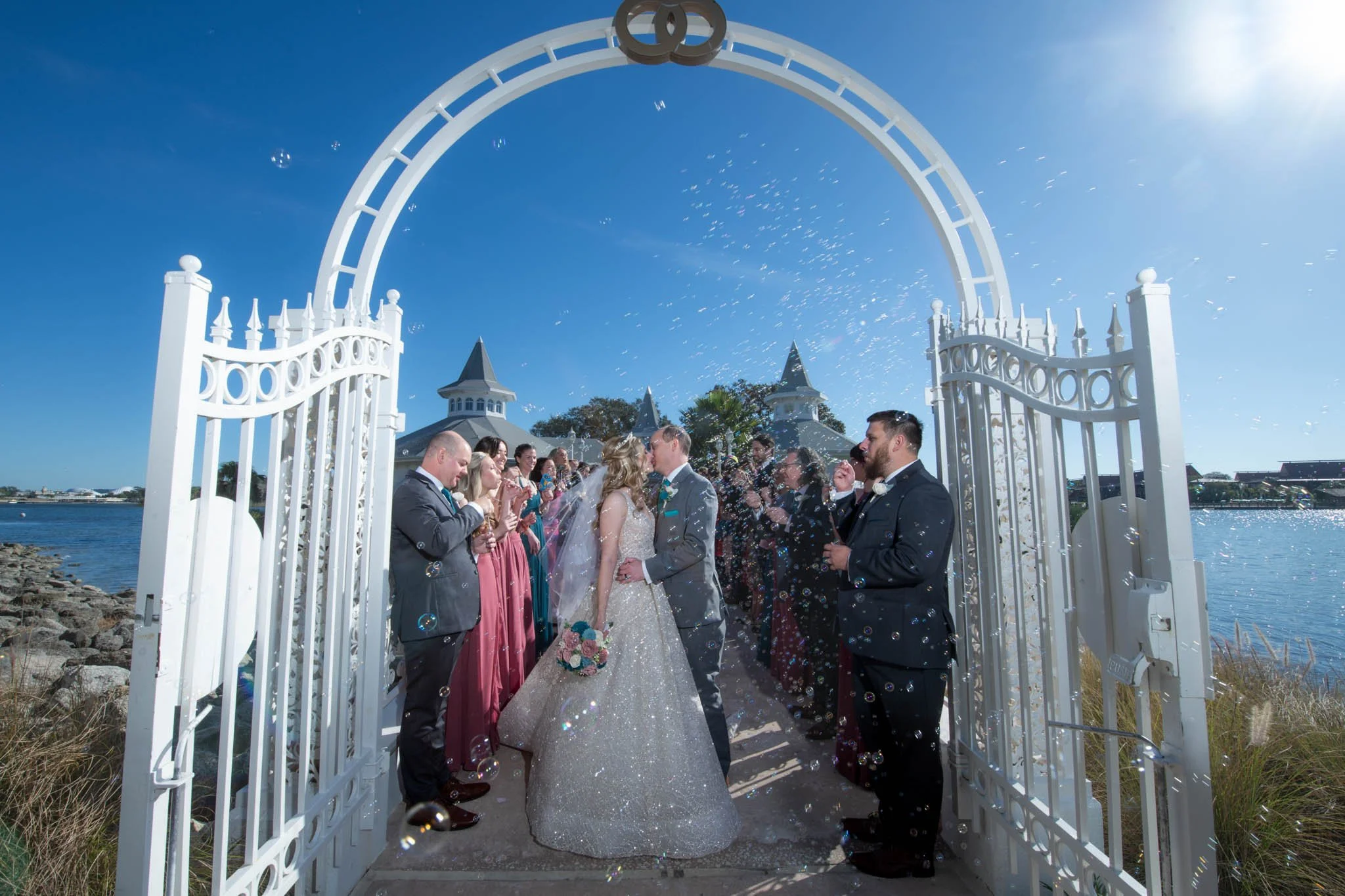 Bride and groom kissing under a white gate during their Disney Wedding Pavilion wedding, with guests celebrating and blowing bubbles on a sunny day by the water.
