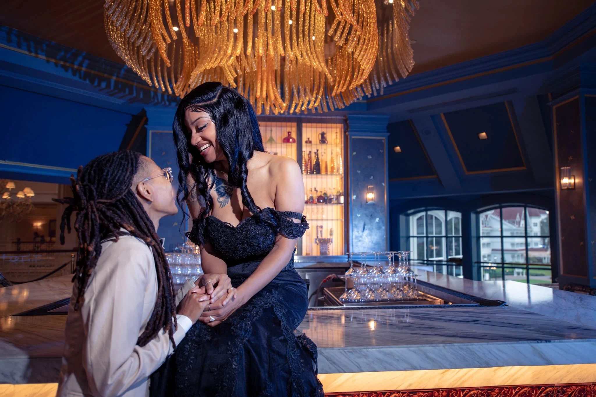 Two women in an elegant restaurant, one with long dreadlocks and glasses, wearing a beige suit, and the other with long black hair, wearing a black off-shoulder dress, holding hands and smiling at each other. A large chandelier hangs above them, and 