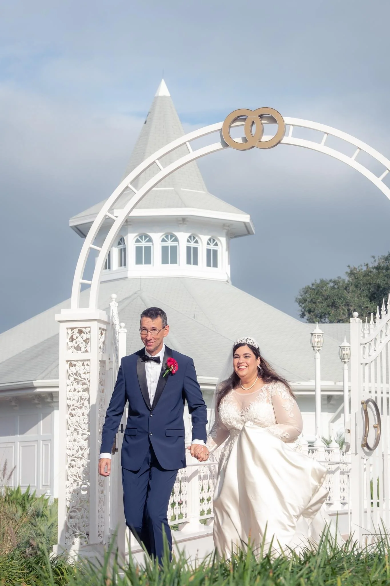 A newlywed couple in wedding attire walking hand in hand outdoors in front of a white decorative arch with wedding rings , with Disney's Wedding Pavilion in the background.