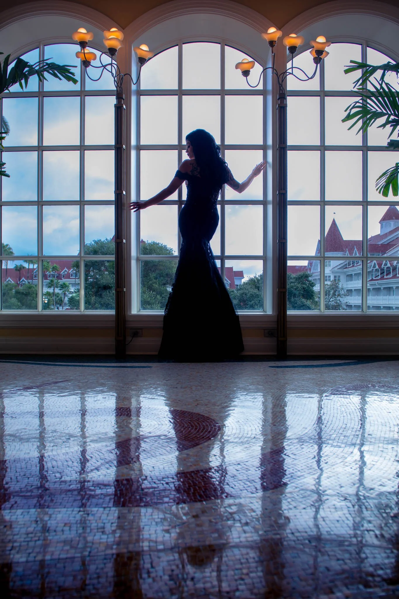 Silhouette of a woman in a long dress standing in front of large window with a scenic view of blue sky, trees, and red-roofed buildings, with indoor plants and an ornate chandelier overhead.