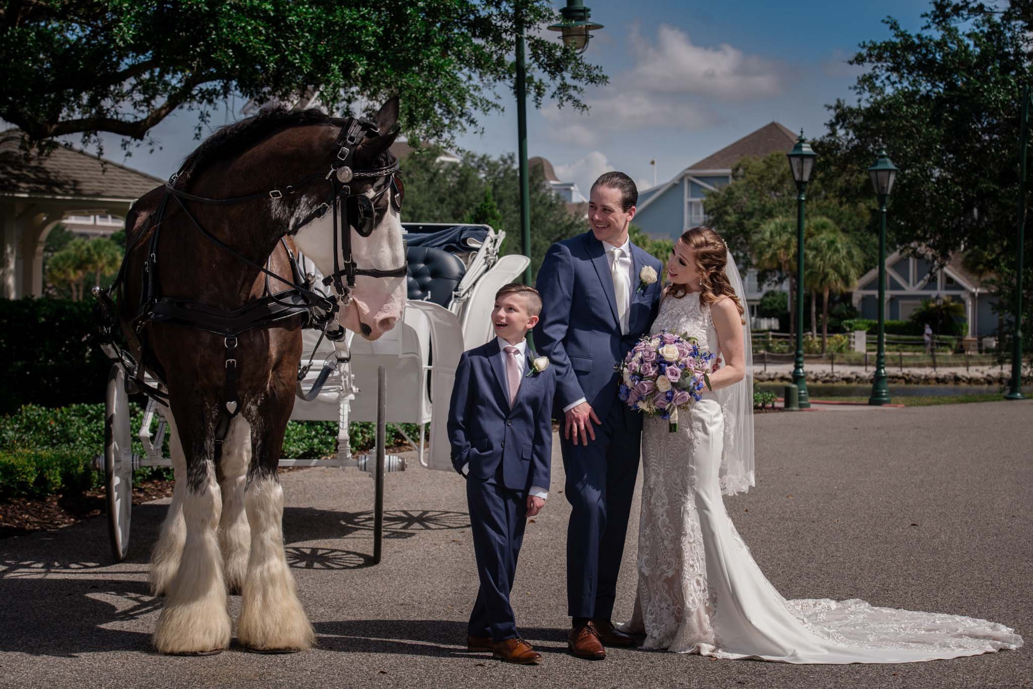 A bride and groom with a young boy, standing outdoors near a white carriage and a large horse. The bride is holding a bouquet of purple and white flowers. The scene is sunny with a blue sky and some trees and houses in the background.