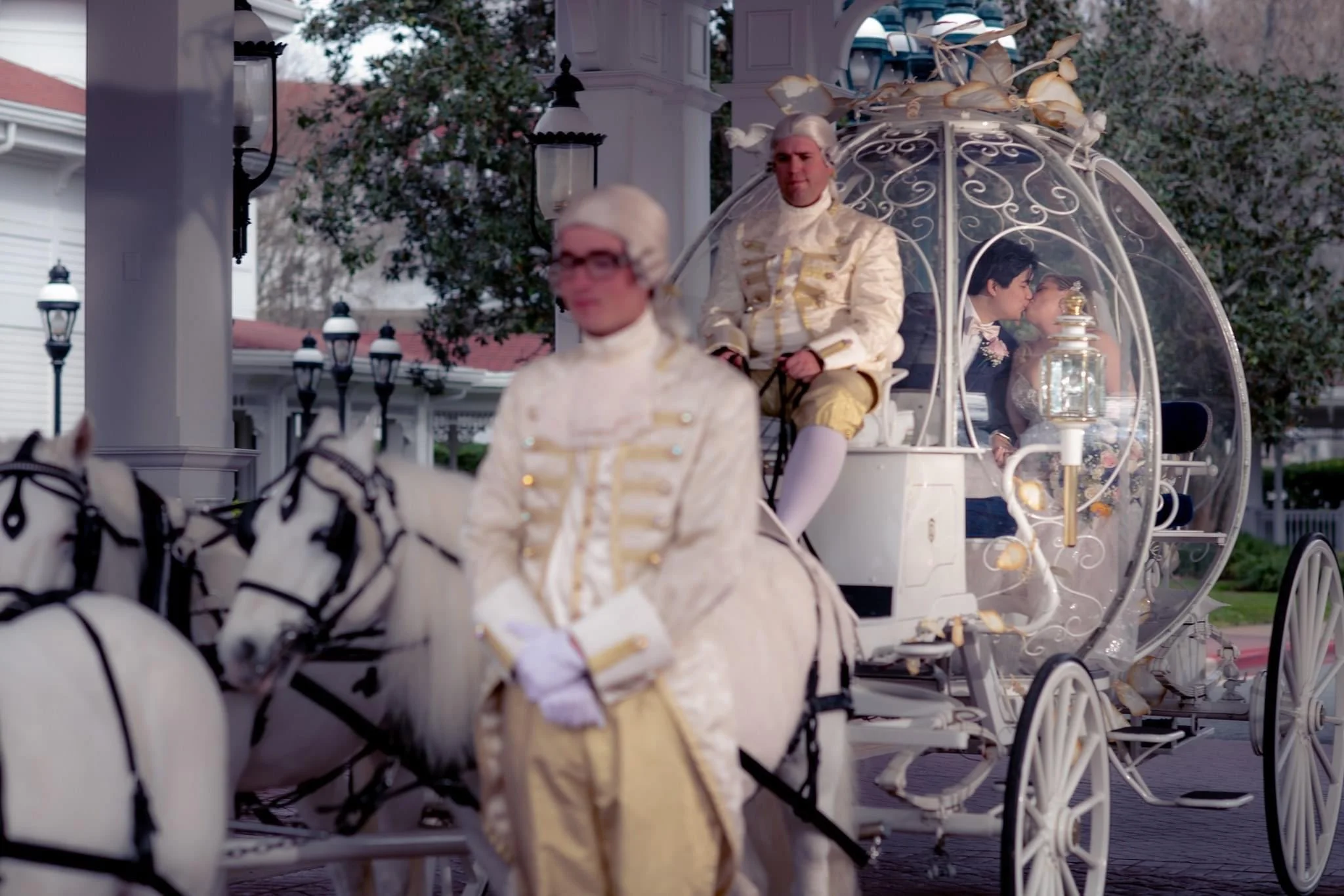 A white horse-drawn Cinderella's carriage carrying a couple kissing inside, with a coach driver and a man in historic costumes leading the horses in front of a white house.