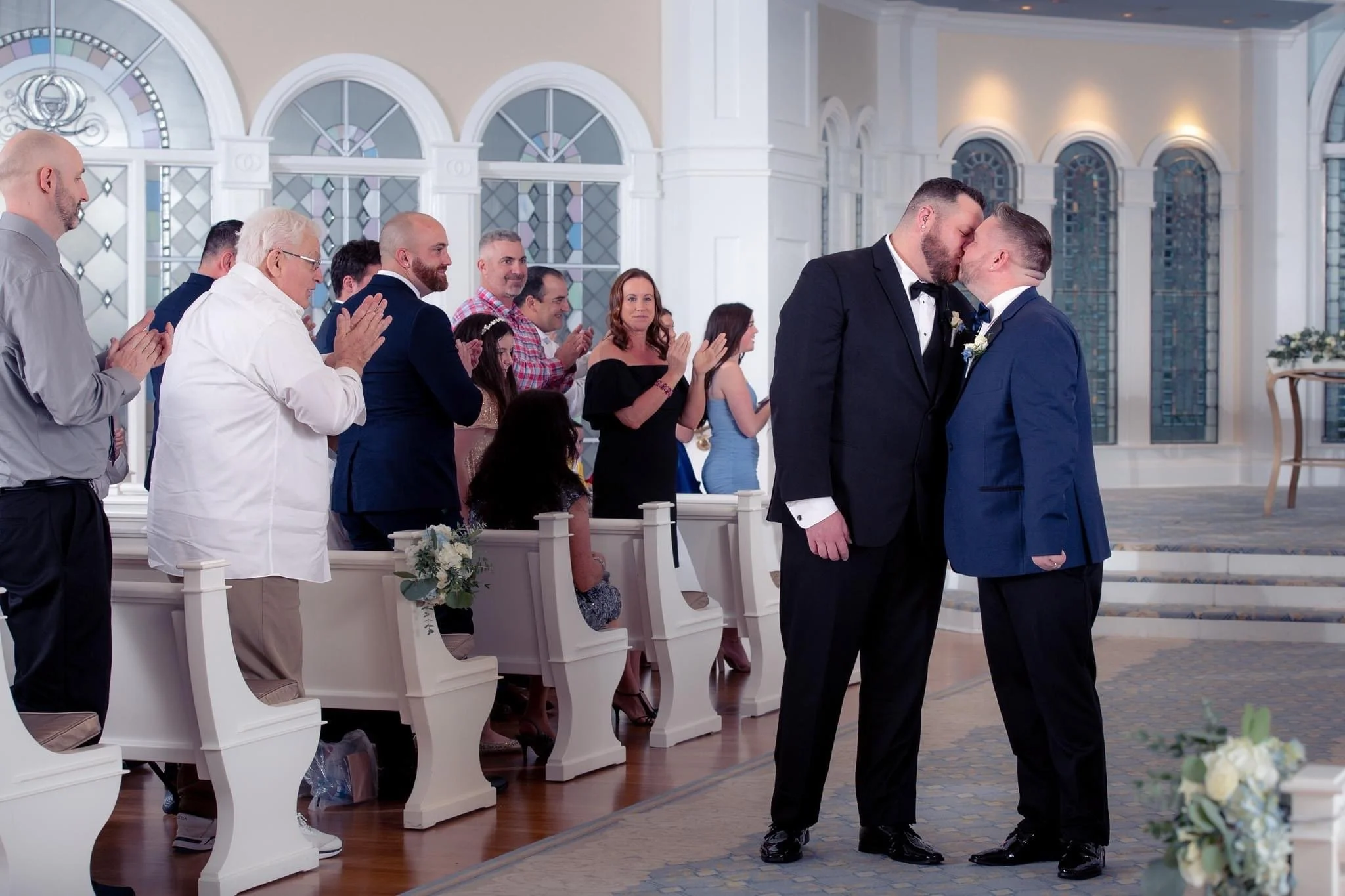 A same-sex couple, both in tuxedos, kiss in Disney's Wedding Pavilion during a wedding ceremony while guests applaud and watch.