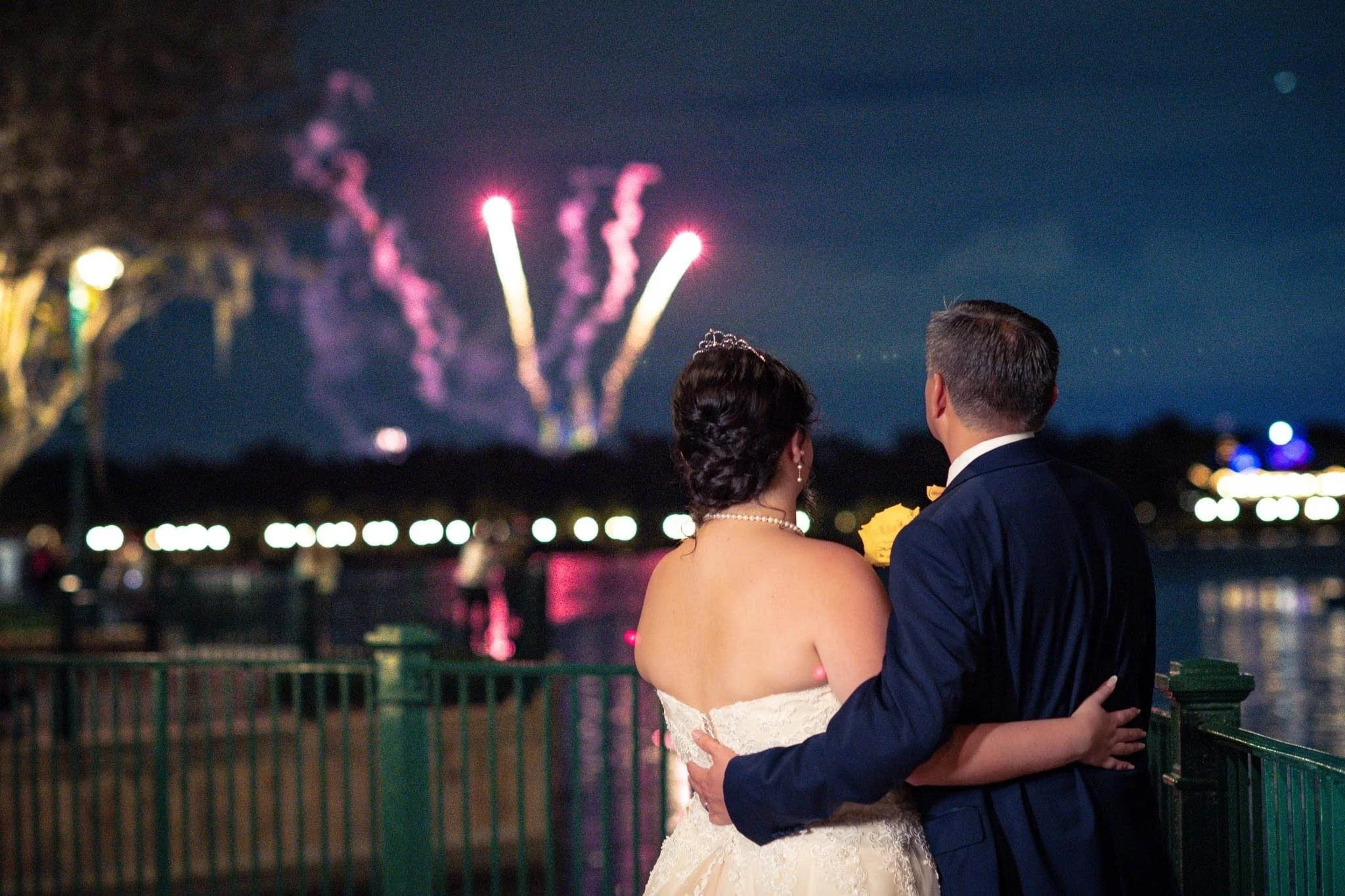 A newlywed couple with their backs to the camera, watching Disney fireworks over a river at night.
