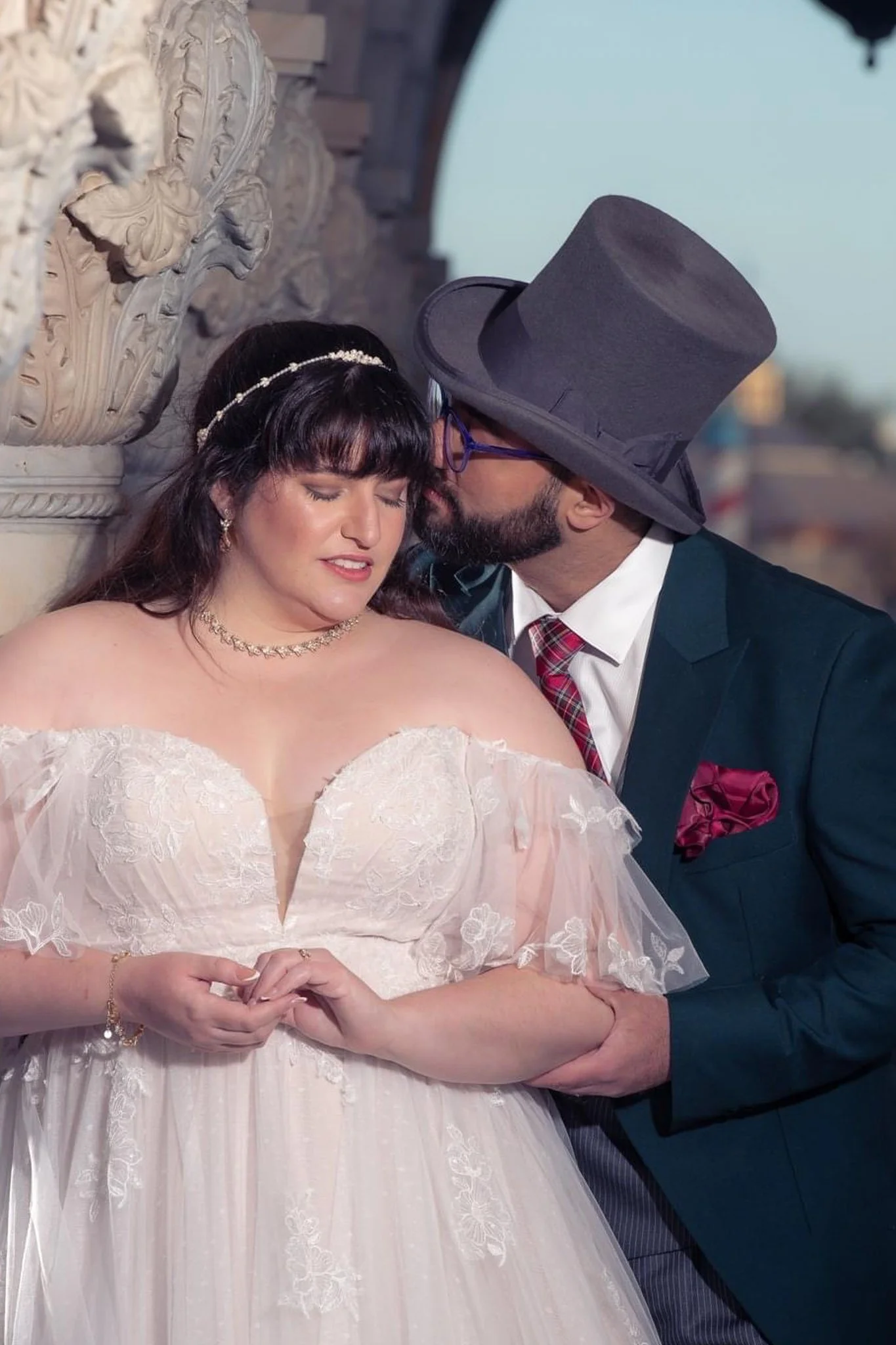 A bride and groom sharing an intimate moment on their wedding day. The bride is wearing a white dress with lace details, and the groom is dressed in a dark suit with a large gray hat and a red tie. They are standing close to a decorative stone wall.