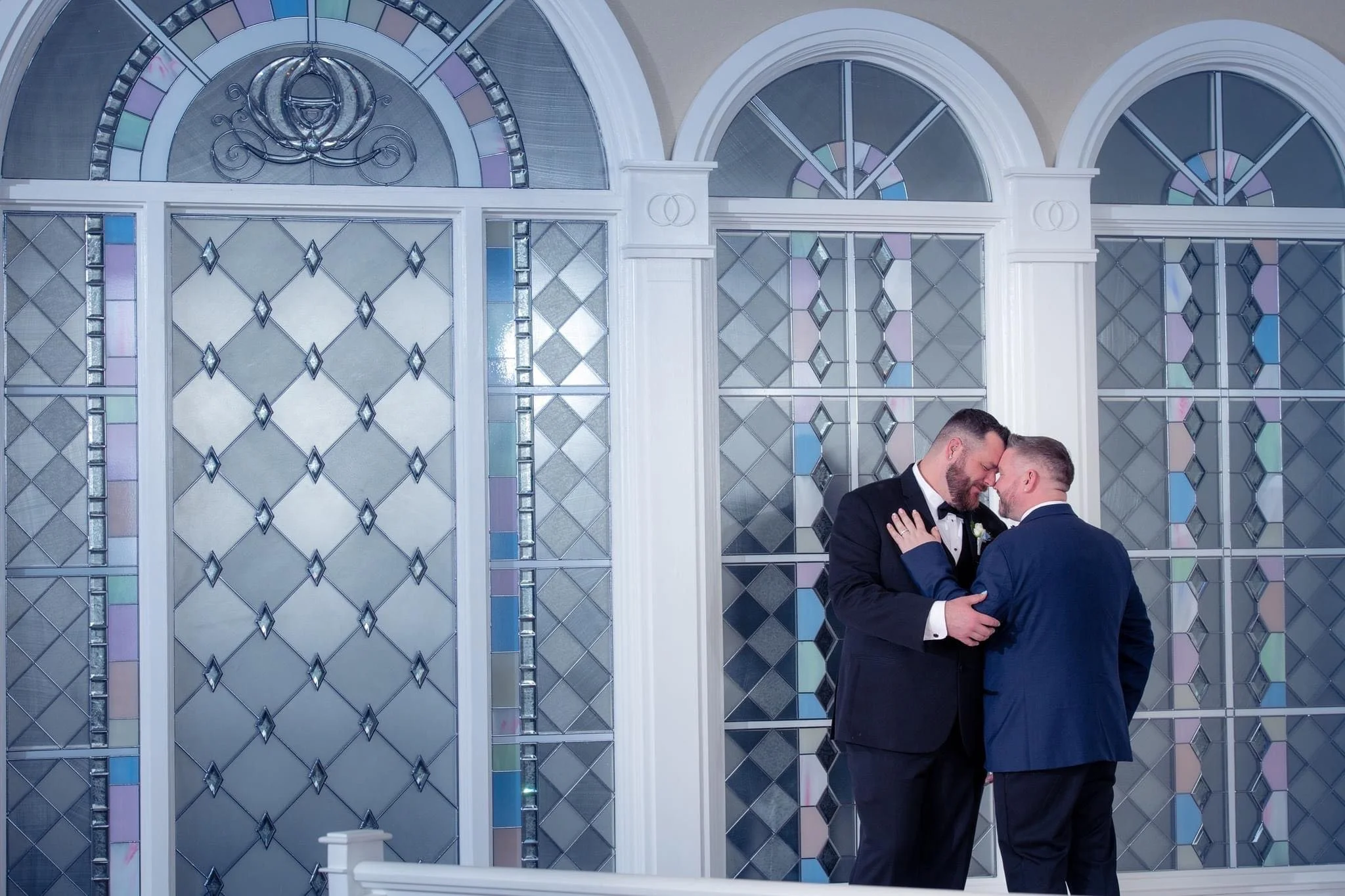 Two men in tuxedos sharing an intimate moment with foreheads touching, standing in front of stained glass windows.
