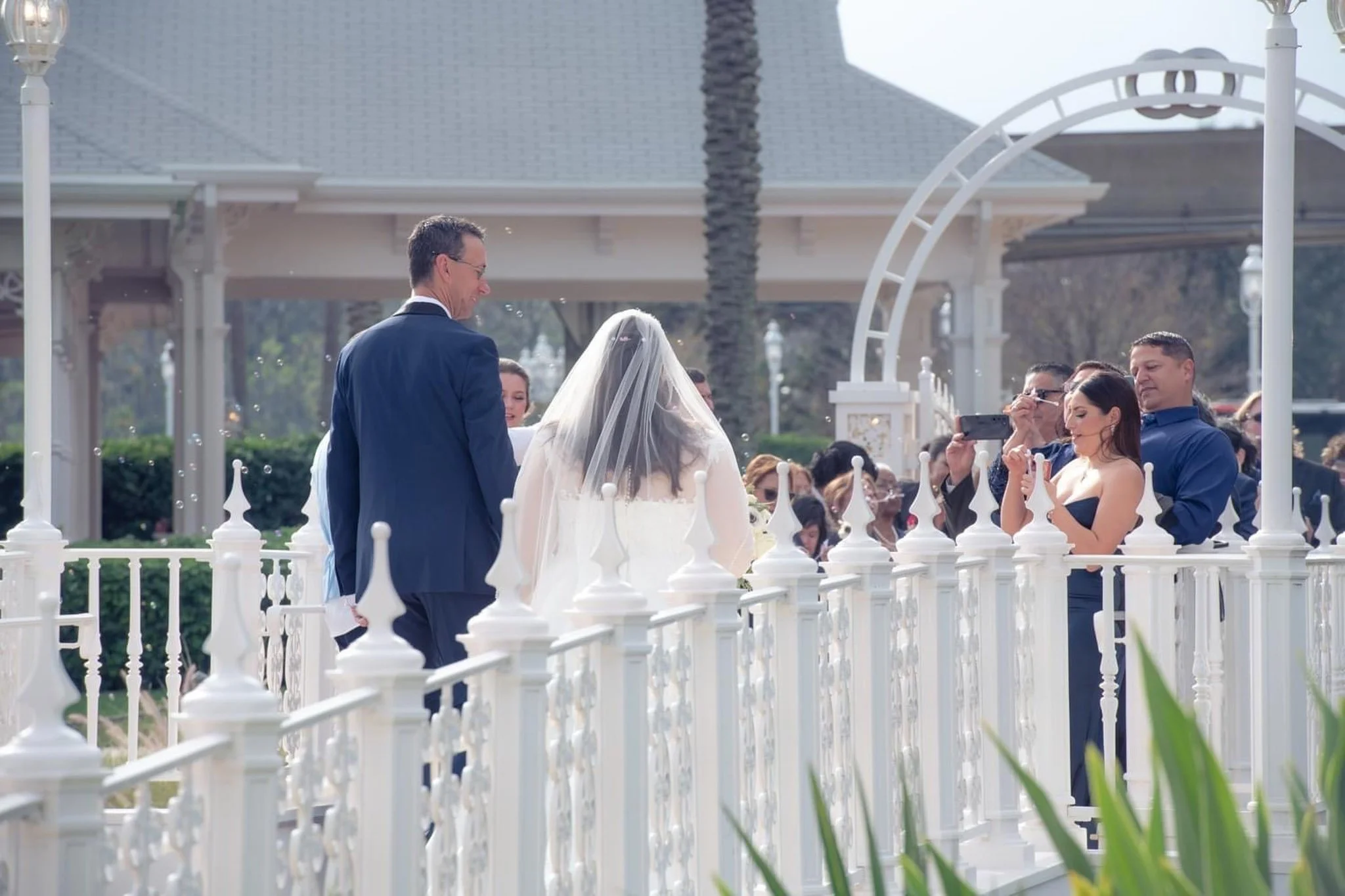 A wedding ceremony outdoors with a bride in a white wedding dress and veil, a groom in a navy suit, and several guests taking photos.