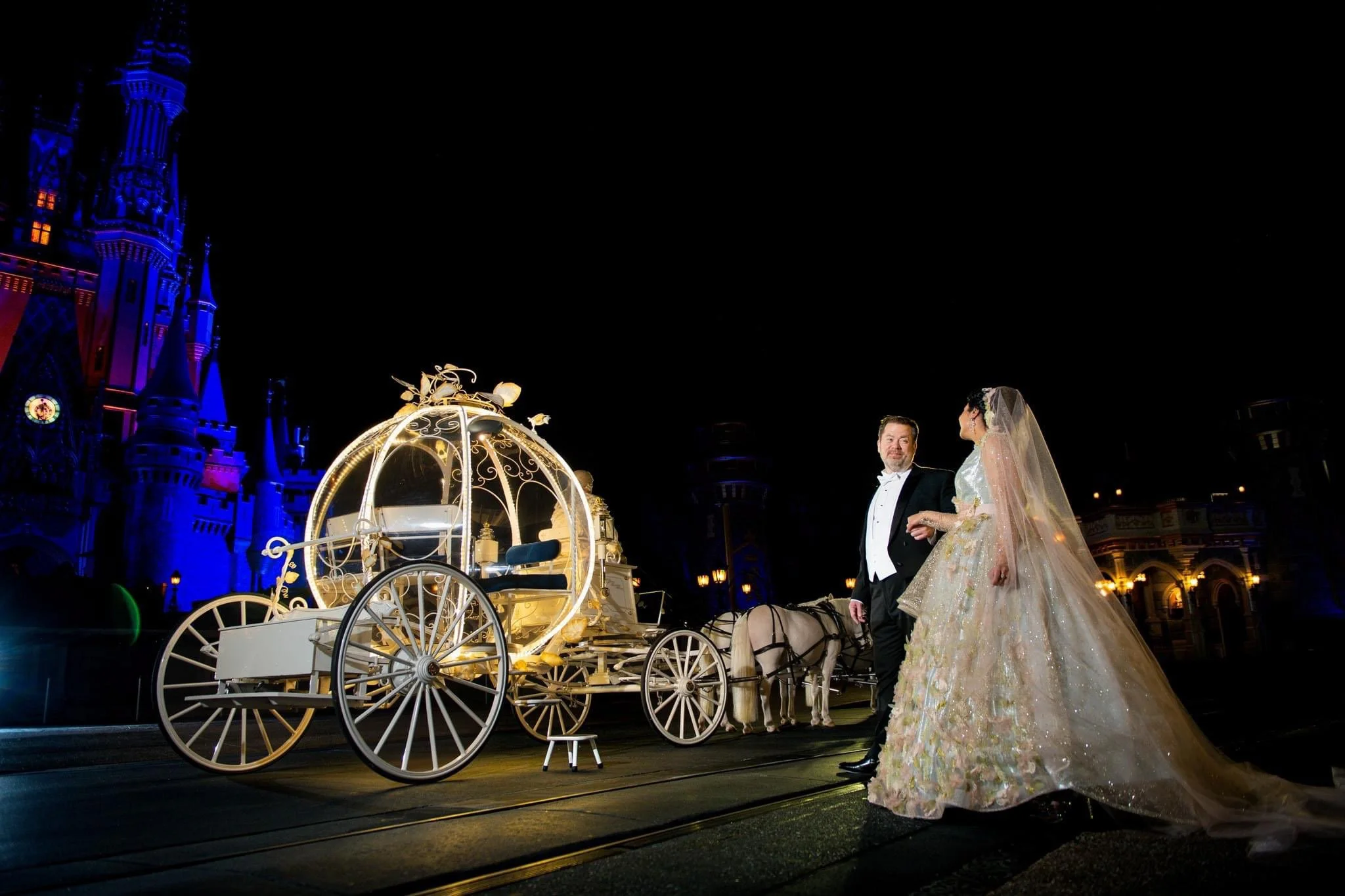 A bride and groom in wedding attire standing near Cinderella's carriage illuminated at night, with Magic Kingdom castle in the background.