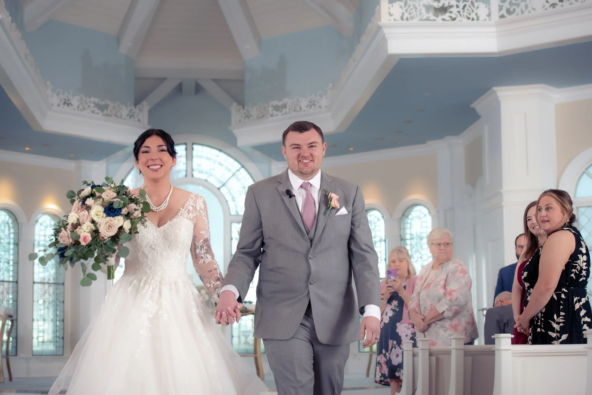 A bride in a white wedding gown holding a large bouquet and a groom in a gray suit holding hands, walking down the aisle inside Disney's wedding pavilion with guests watching.