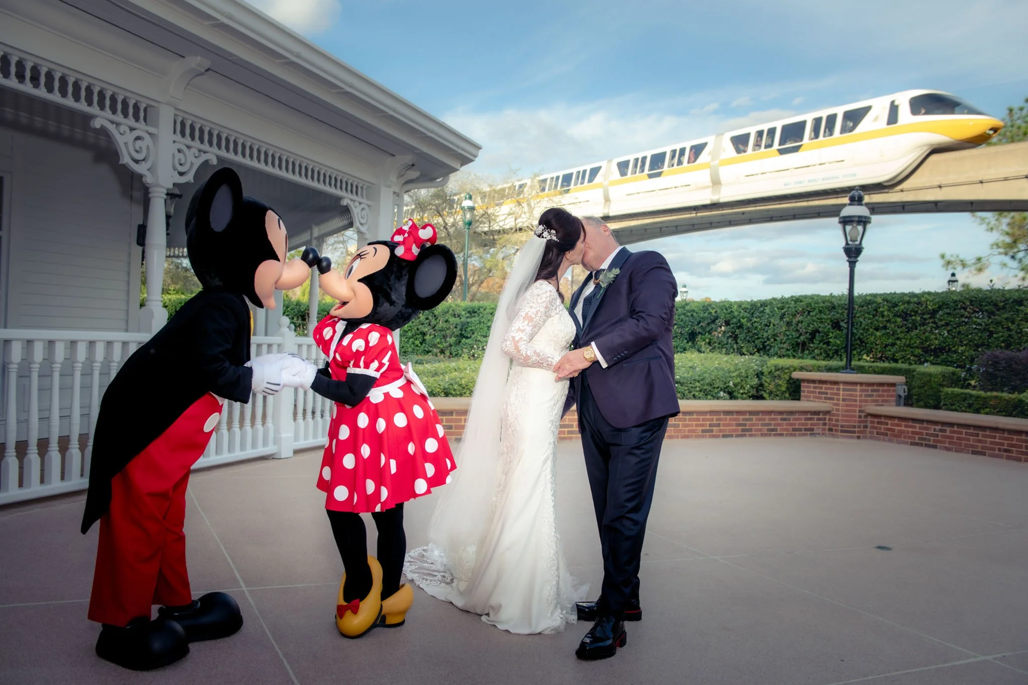 A bride and groom kiss on their wedding day with Mickey and Minnie Mouse characters holding hands nearby, in front of a monorail train passing on elevated tracks at White Hall at The Grand Floridian.