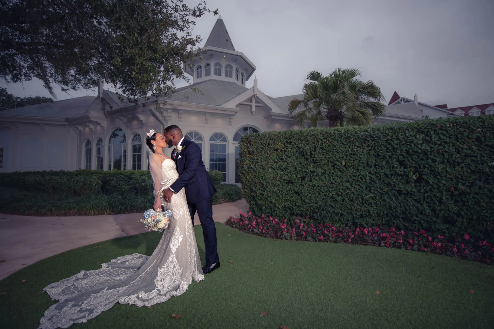 A bride and groom dressed in wedding attire, sharing a kiss outdoors in front of a large, white, Victorian-style house with a tower, surrounded by greenery and flower beds at Disney's Wedding Pavilion.