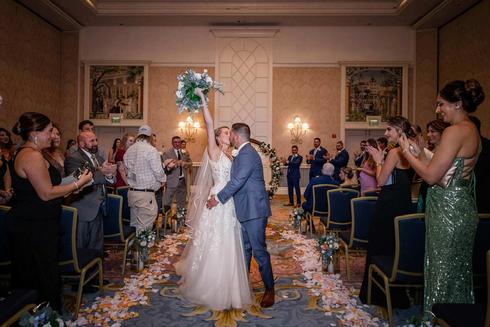 A bride and groom share a kiss in a wedding ceremony, with the bride holding a bouquet up in the air. Guests stand and applaud along the aisle, which is decorated with flower petals and floral arrangements. The ceremony is held in an elegant indoor v