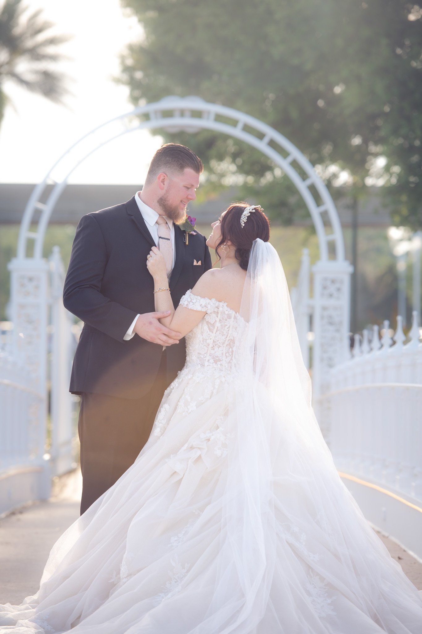 A bride and groom sharing a tender moment outdoors during their wedding, with a white decorative arch and trees in the background at Disney's Wedding Pavilion.