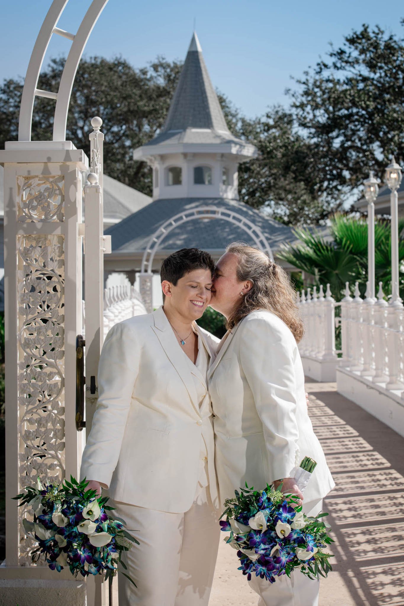 Two people in white suits, one kissing the other's forehead, holding bouquets, standing in front of a white ornate gate and Disney's Wedding pavilion with a conical steeple, outdoors on a sunny day.