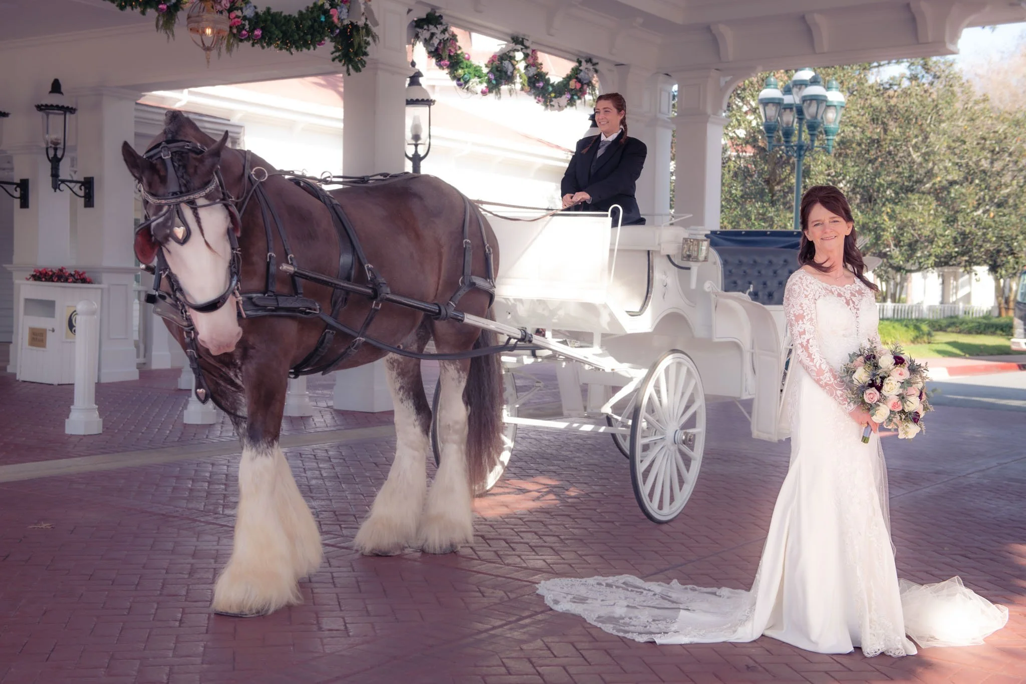 A bride in a white wedding dress holding a bouquet stands in front of a white horse-drawn carriage, with a woman sitting in the carriage. The scene is outdoors with trees and a brick paved area at The Grand Floridian.