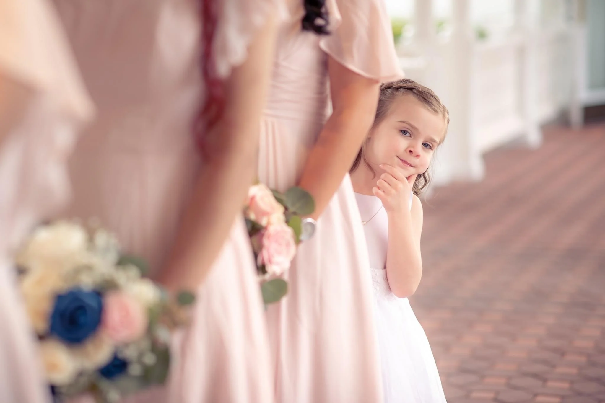 A young girl in a white dress looking thoughtfully with her hand on her chin while standing among women dressed in pink formal dresses holding bouquets of flowers, in a bright, elegant setting.