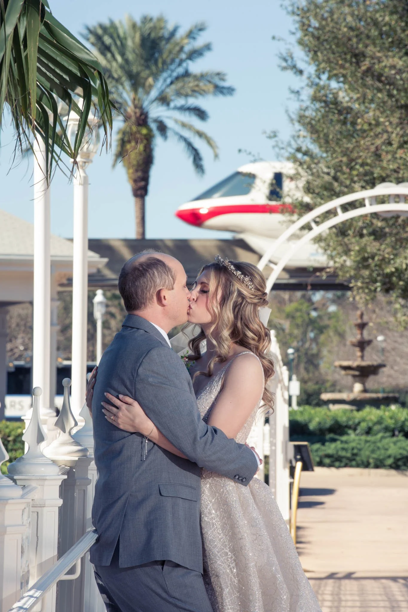 A newlywed couple sharing a kiss outdoors, with a Disney monorail in the background and palm trees visible.
