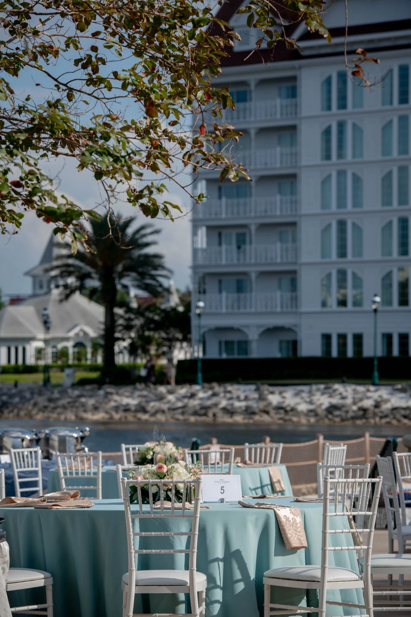 Outdoor event setup with round tables covered in light blue tablecloths, decorated with floral centerpieces, and surrounded by white chairs. In the background, there is a water body, a tall palm tree, and a large white building, suggesting a seaside 