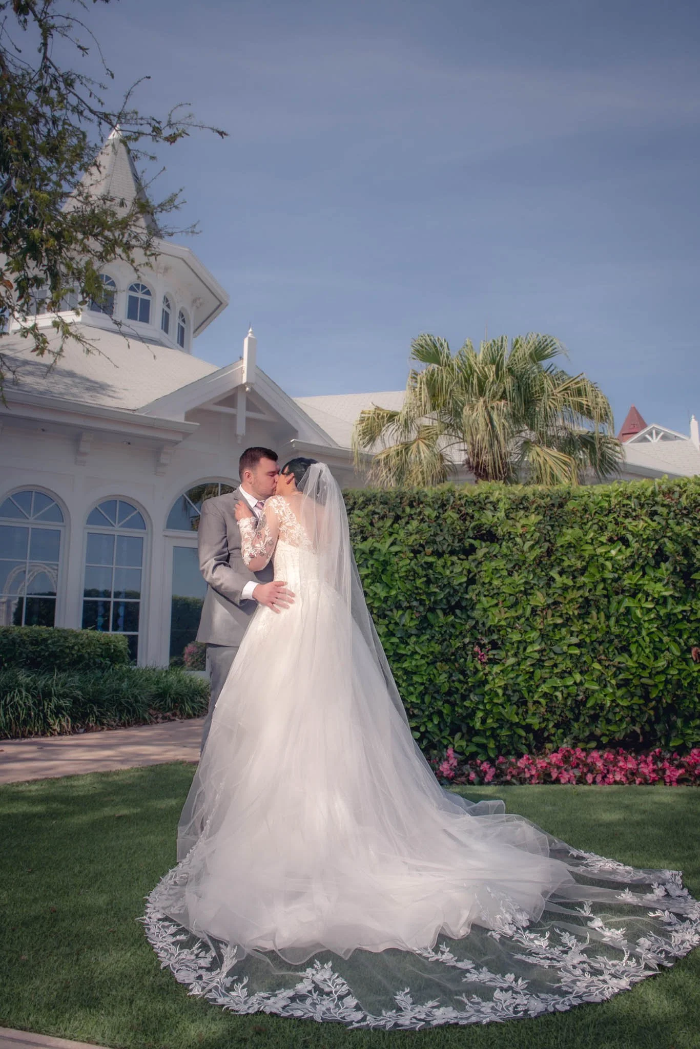 A bride and groom share a kiss outside Disney's Wedding Pavilion, with lush greenery and pink flowers in the background.