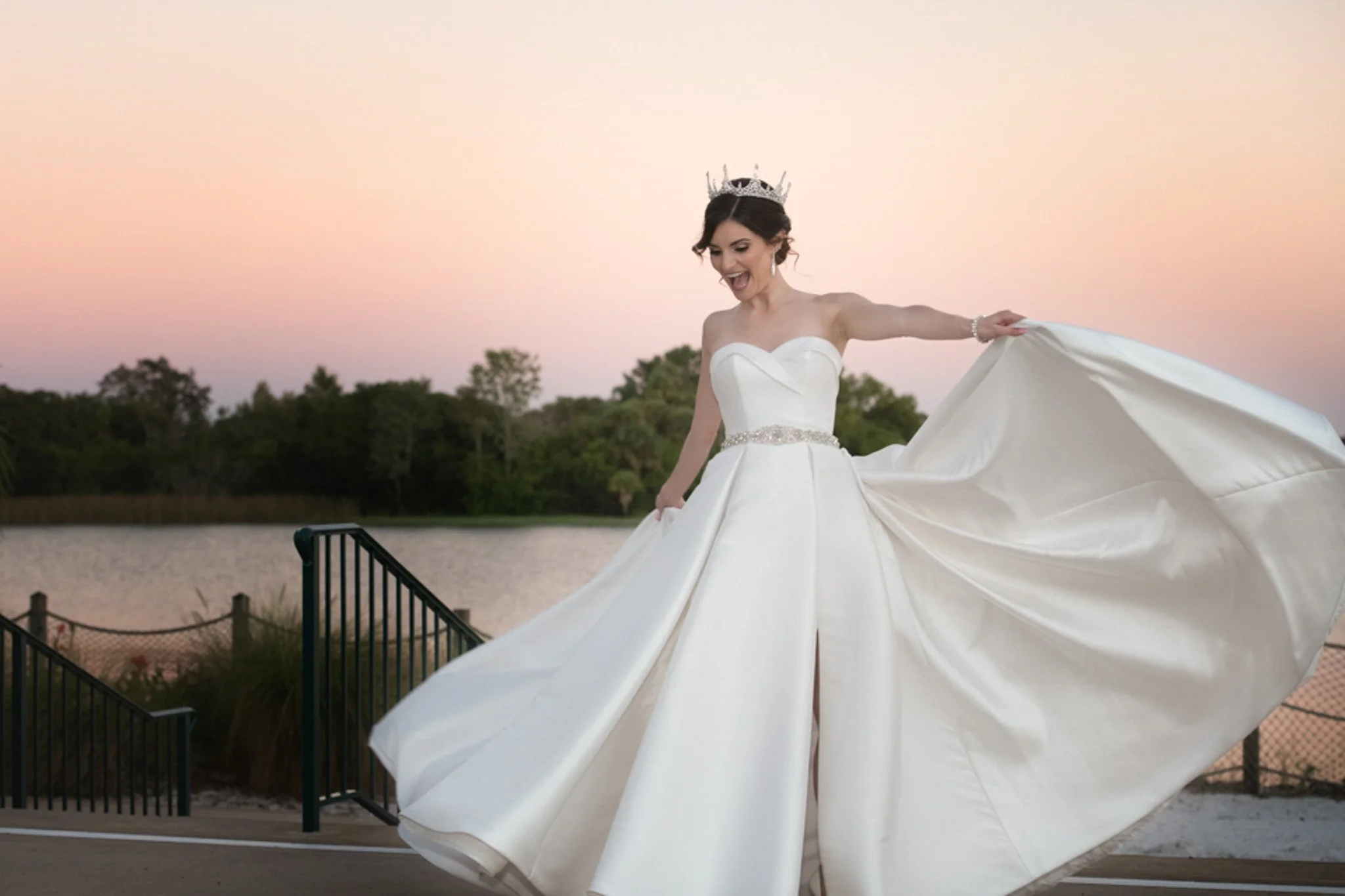 A bride in a strapless white wedding gown with a full skirt and a beaded belt, standing outdoors near a body of water at sunset, smiling and holding up her dress.