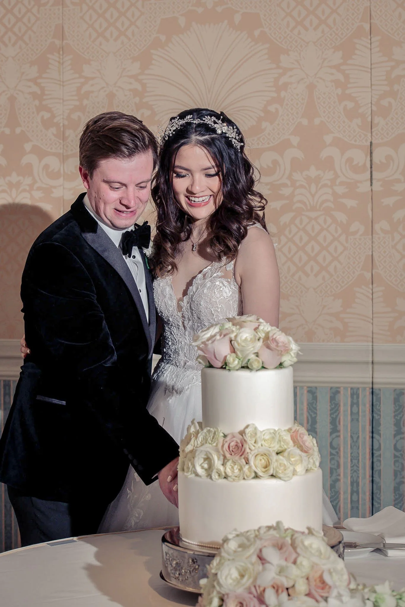 A bride and groom cutting a wedding cake together, with the bride wearing a lace wedding dress and tiara, and the groom in a tuxedo, in an elegant indoor setting.