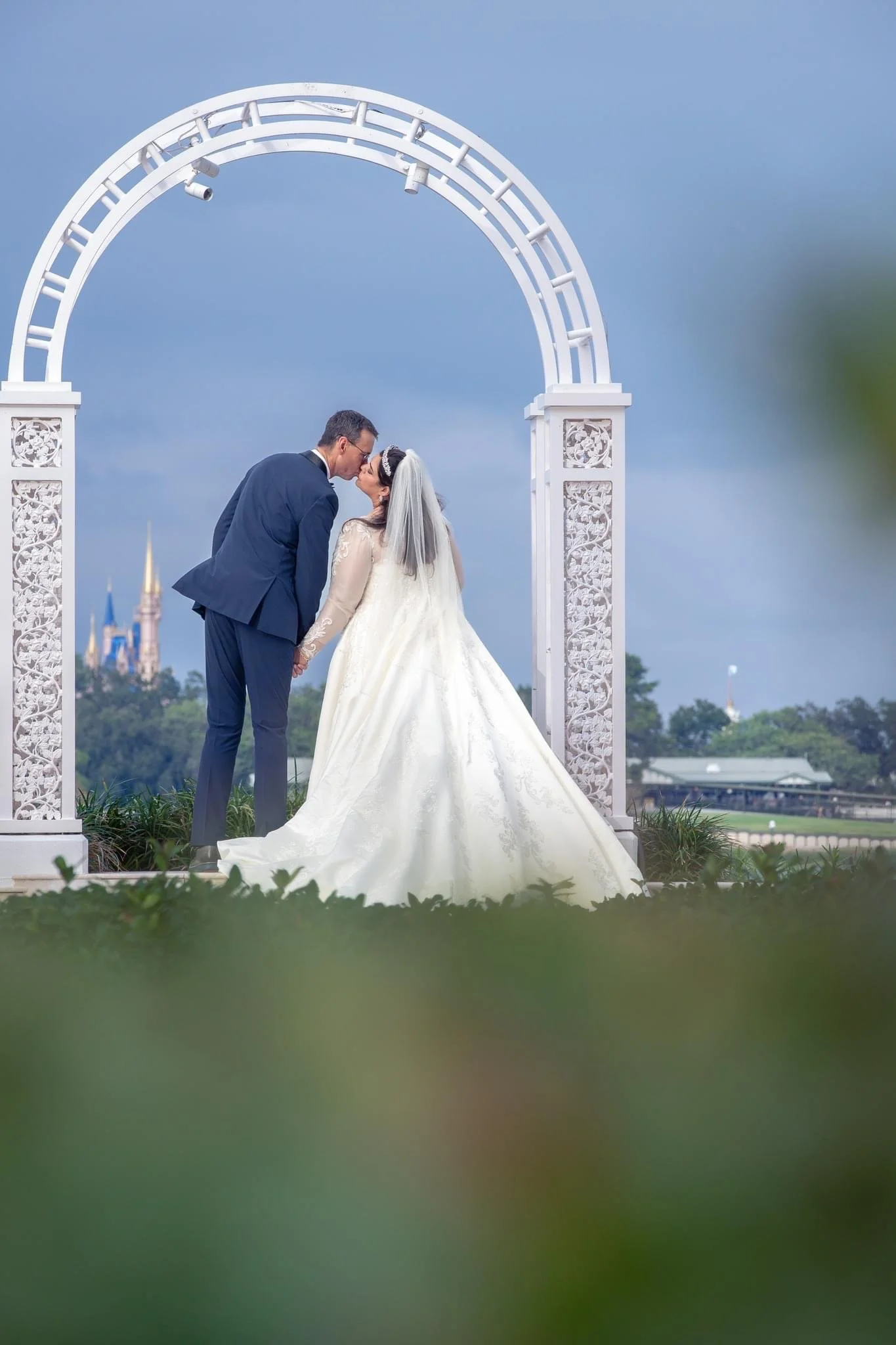 A bride and groom share a kiss on their wedding day under a white decorative arch of Picture Point outdoors, with a castle in the background and cloudy sky.
