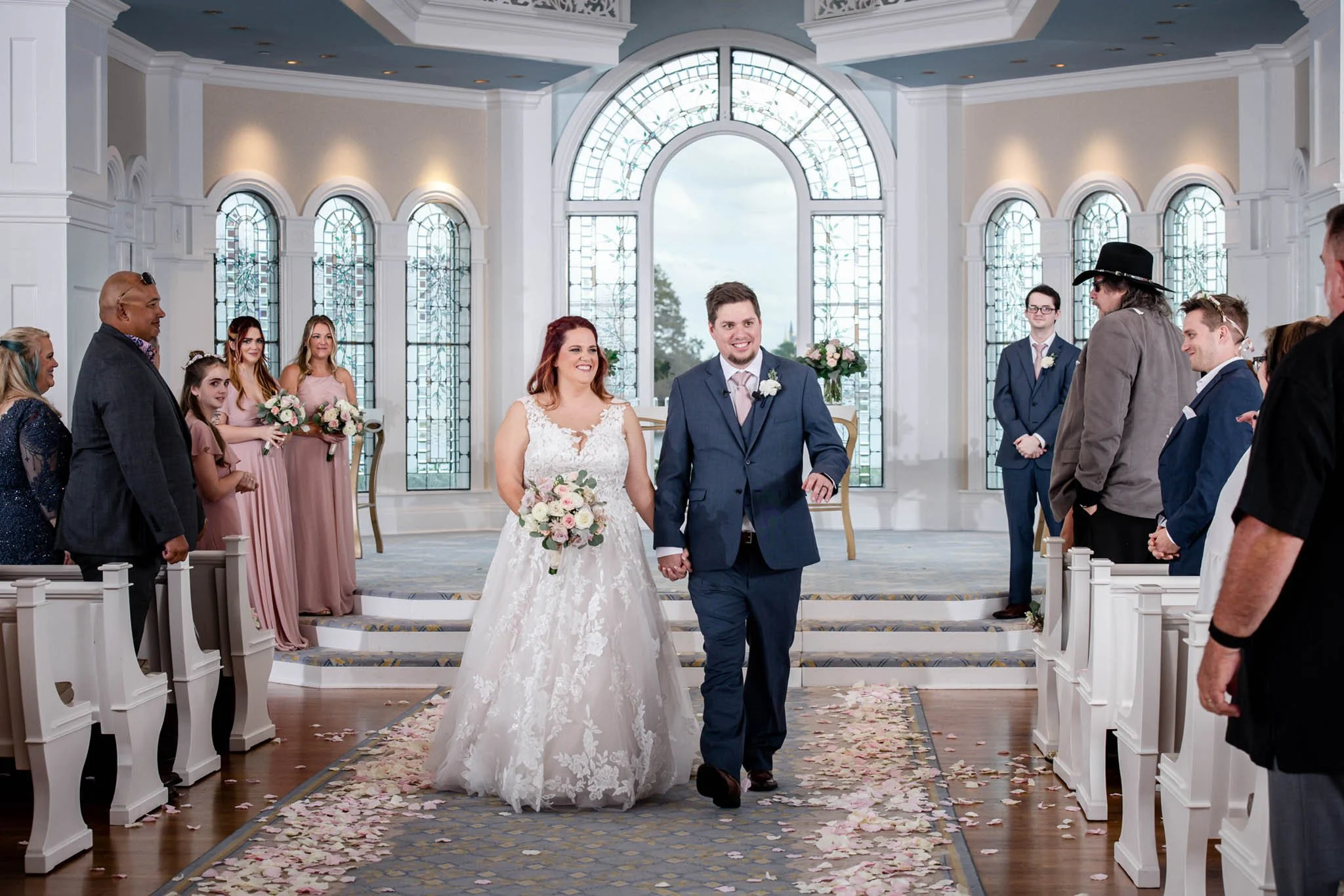 A bride and groom walking down the aisle at a wedding ceremony inside Disney's Wedding Pavilion with stained glass windows, surrounded by friends and family.