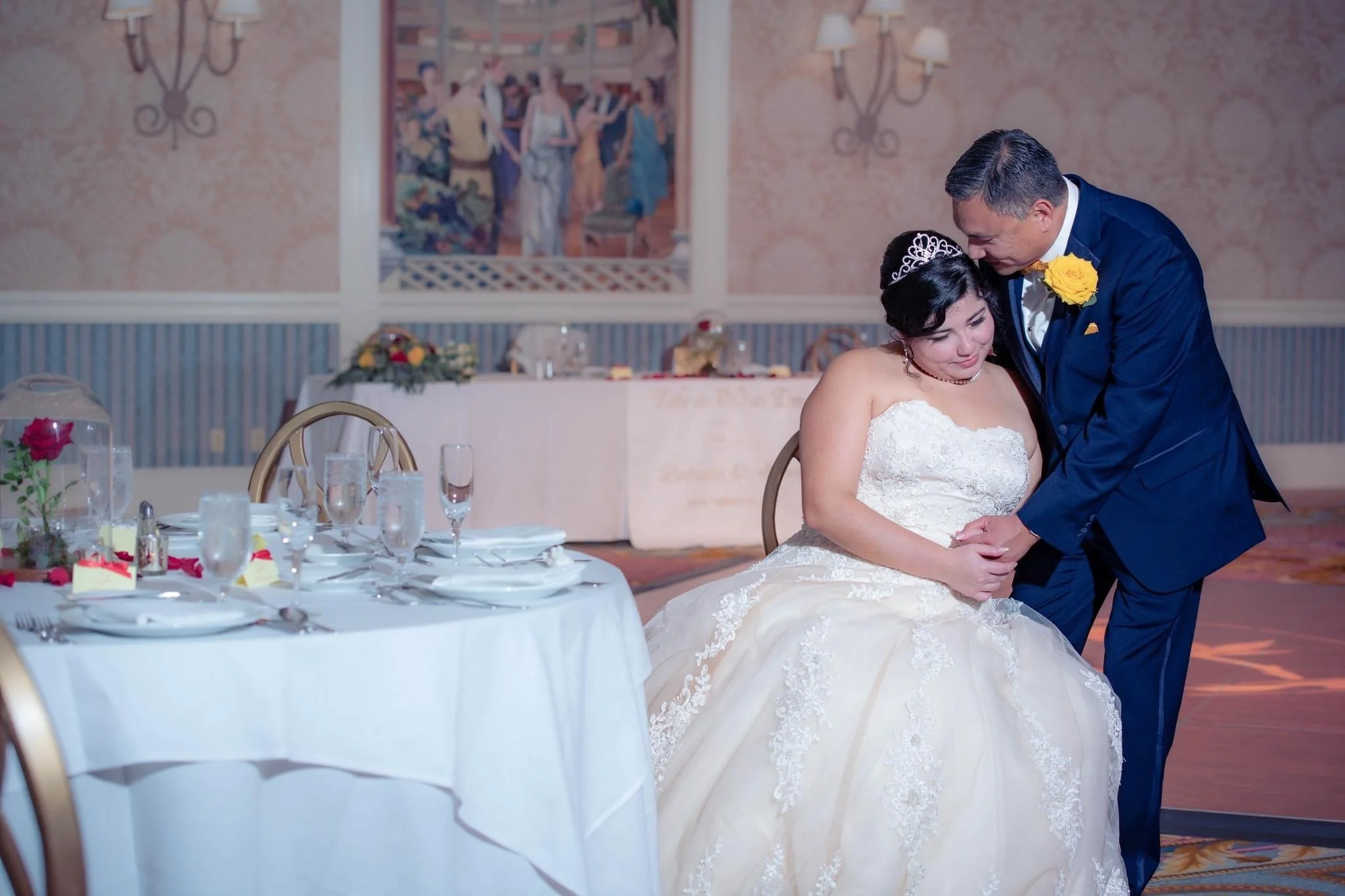 A bride and groom share an intimate moment at their wedding reception; the bride is seated in a white wedding gown, and the groom is bending down to hold her hands and kiss her on the head.