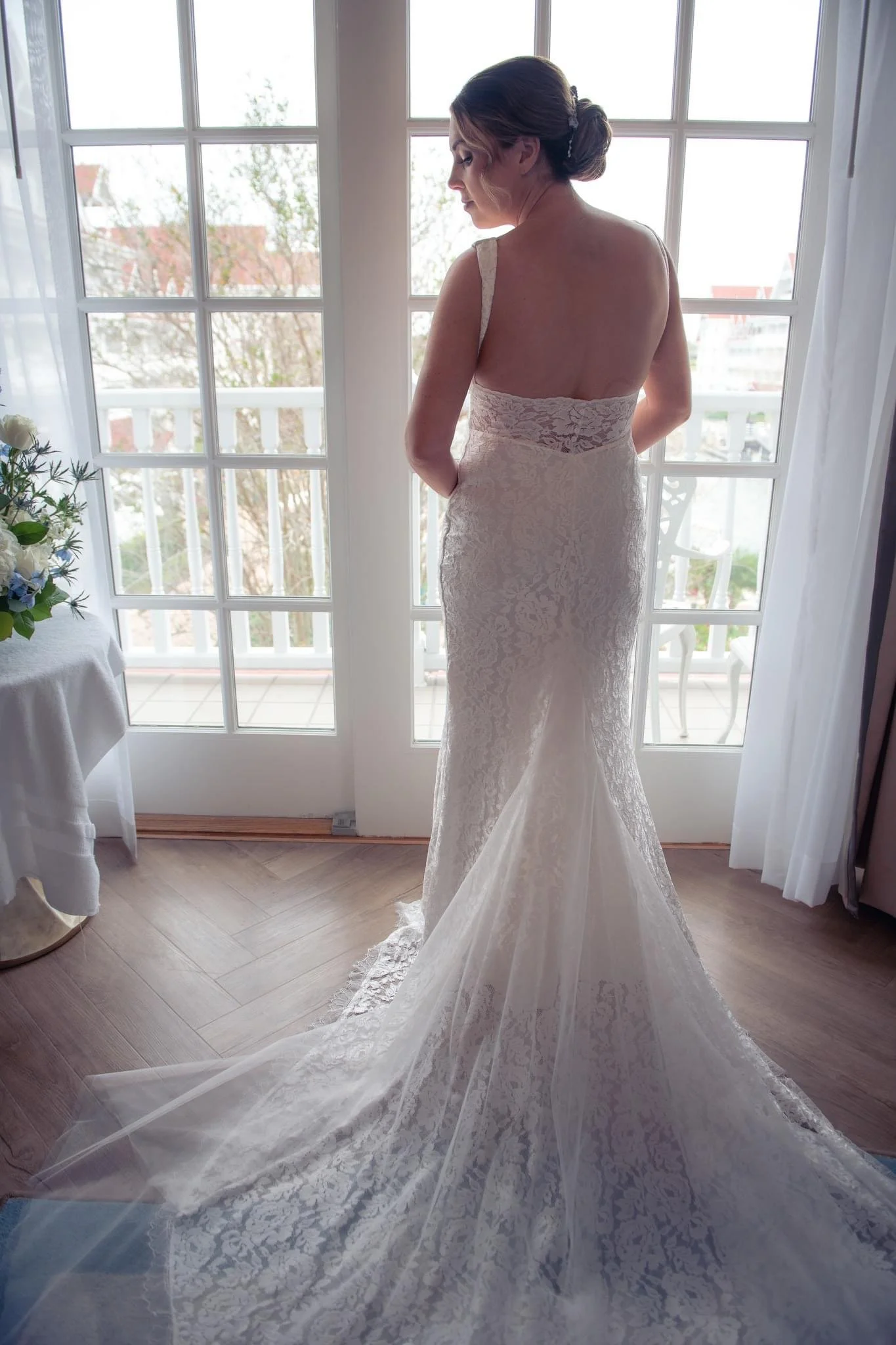 A woman in a white lace wedding dress standing in front of glass doors, looking outside, with a wedding veil trailing on the floor.