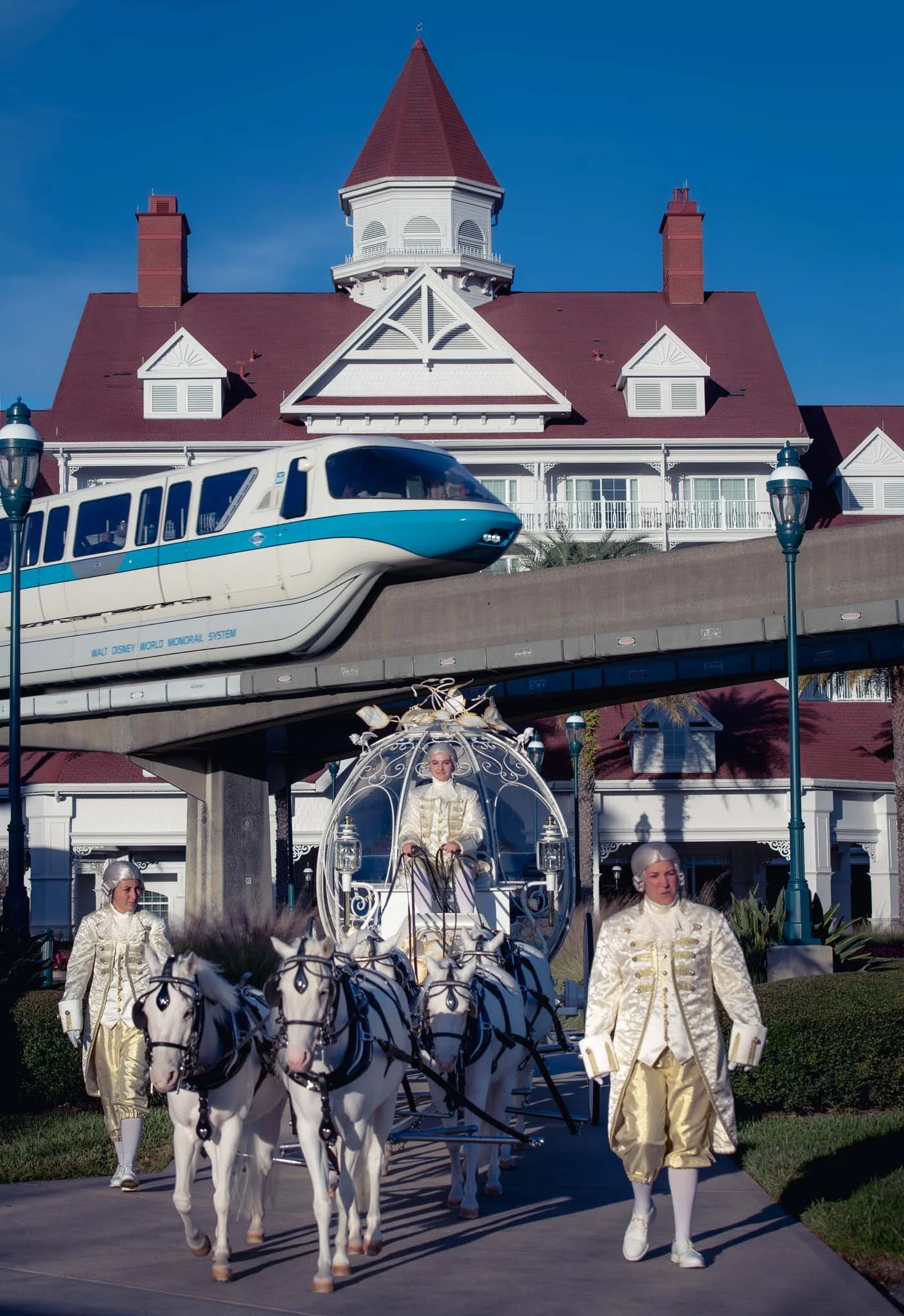 A Bride and Groom ariving in a glass slipper carriage drawn by white horses, with two others in similar costumes walking beside, in front of the Grand Floridian and above a monorail train.