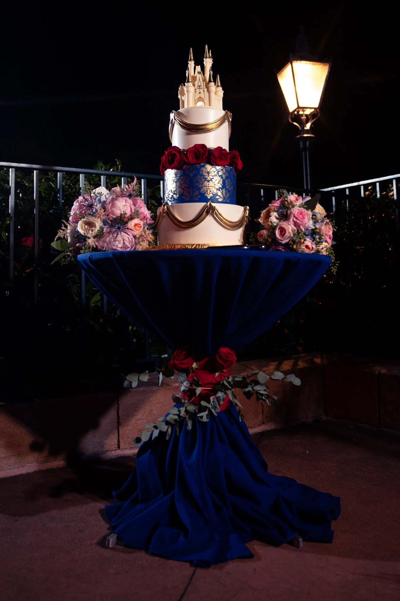 Three-tiered wedding cake decorated with gold accents, red roses, and a castle topper, placed on a table covered with blue fabric, surrounded by pink and purple flowers, at night near a streetlamp.