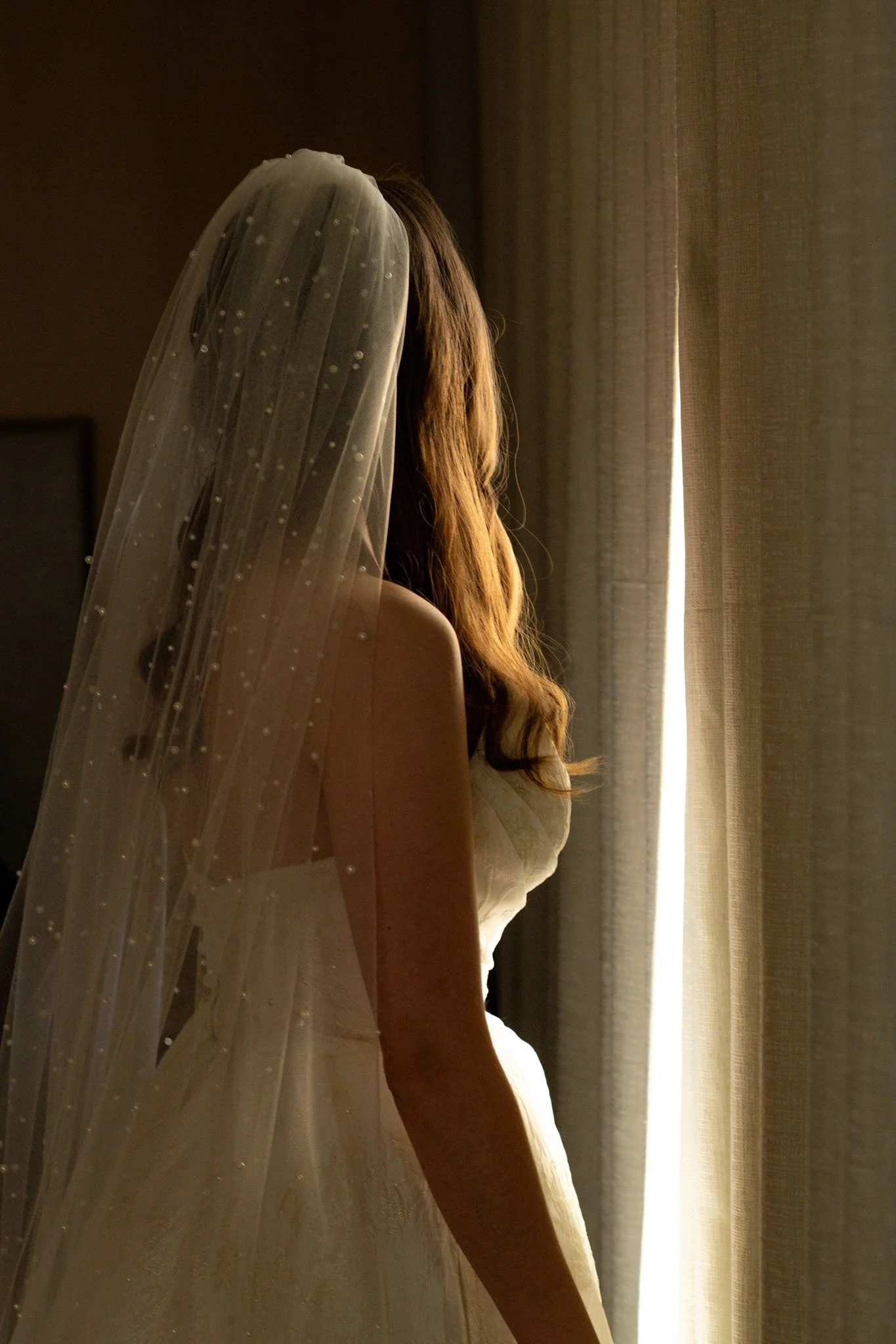 A bride with long hair and a veil gazes out a window, illuminated by sunlight, wearing a strapless wedding dress.