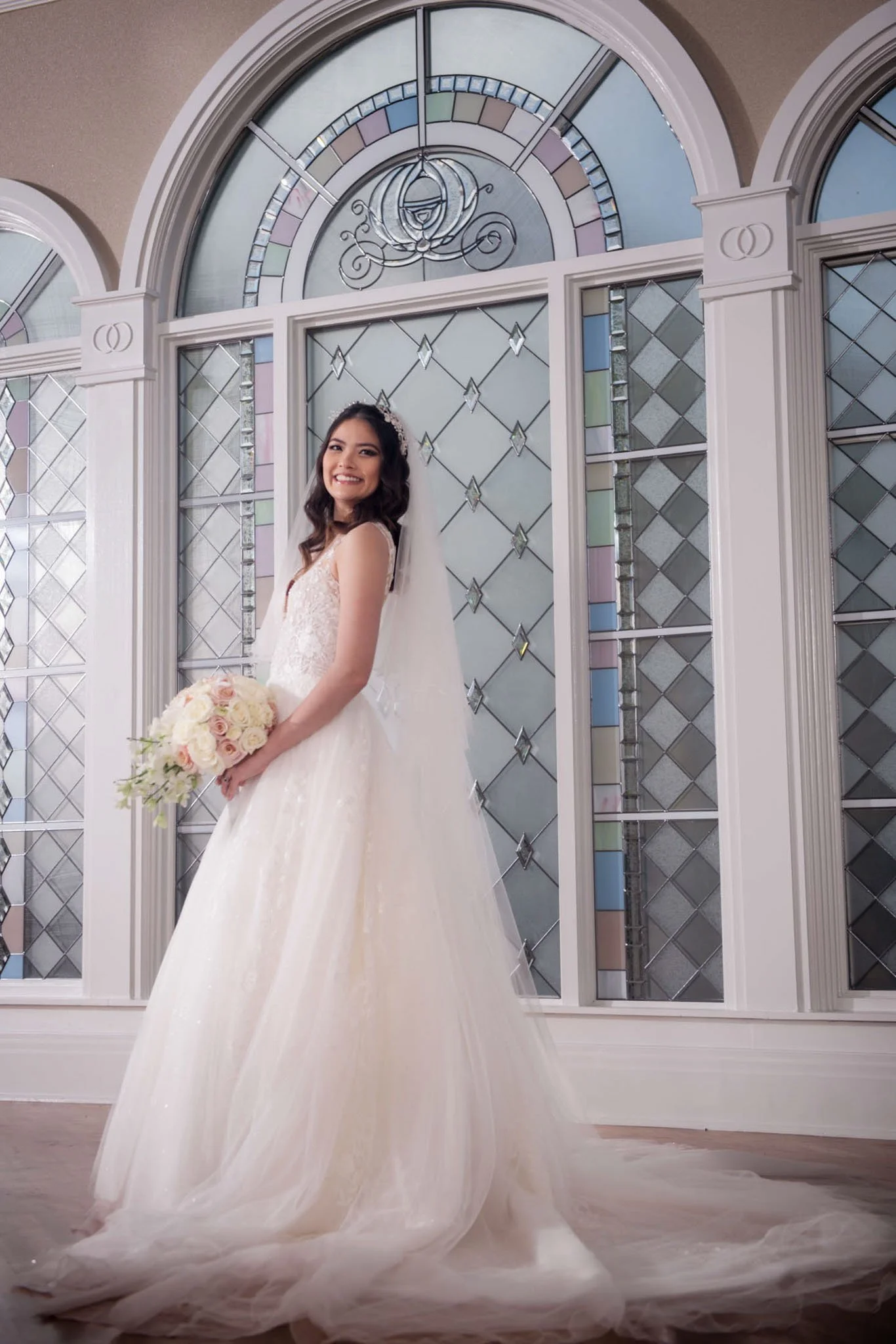 A bride in a white wedding gown holding a bouquet of pink and white roses, standing in front of stained glass windows, smiling at Disney's Wedding Pavilion.