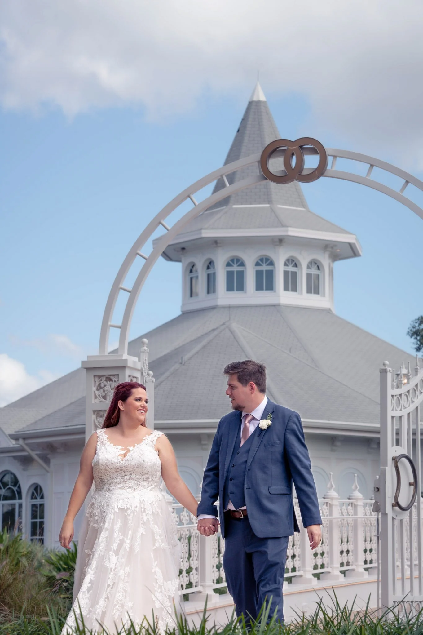 A bride and groom holding hands and walking outside of Disney's Wedding Pavilion with a gray roof and a decorative gate, under a partly cloudy sky.