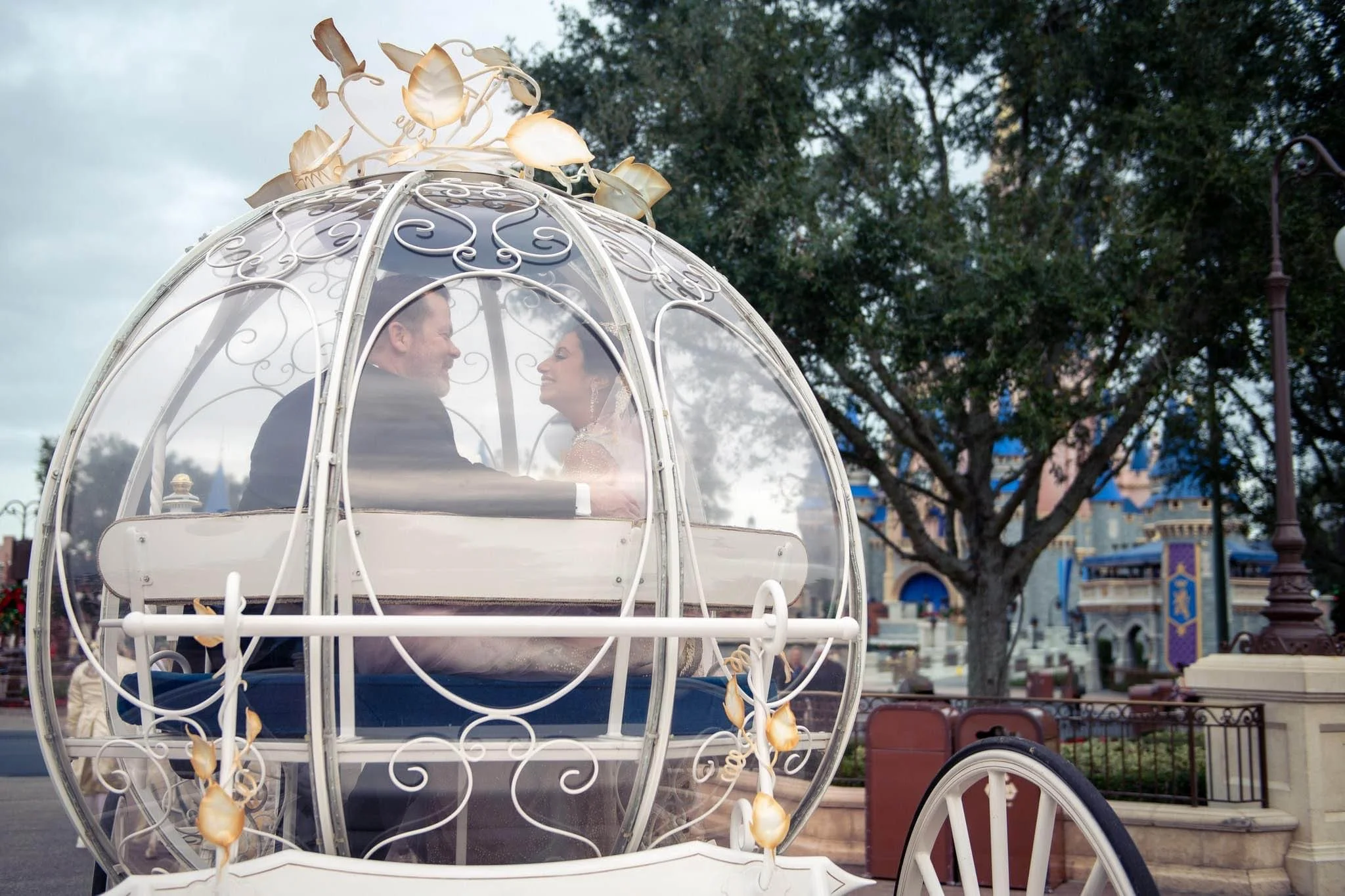 A couple dressed in wedding attire sitting inside a white, decorative glass carriage at a Walt Disney World.