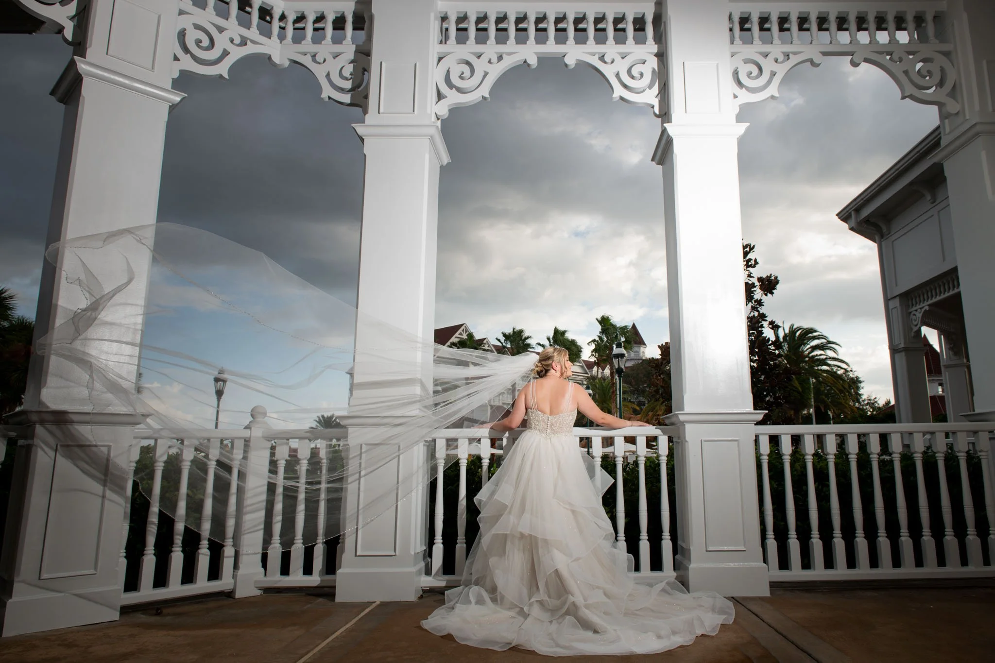 A bride in a white wedding gown with a long veil looking away on a white gazebo at dusk, with dark clouds in the sky and trees in the background of The Grand Floridian