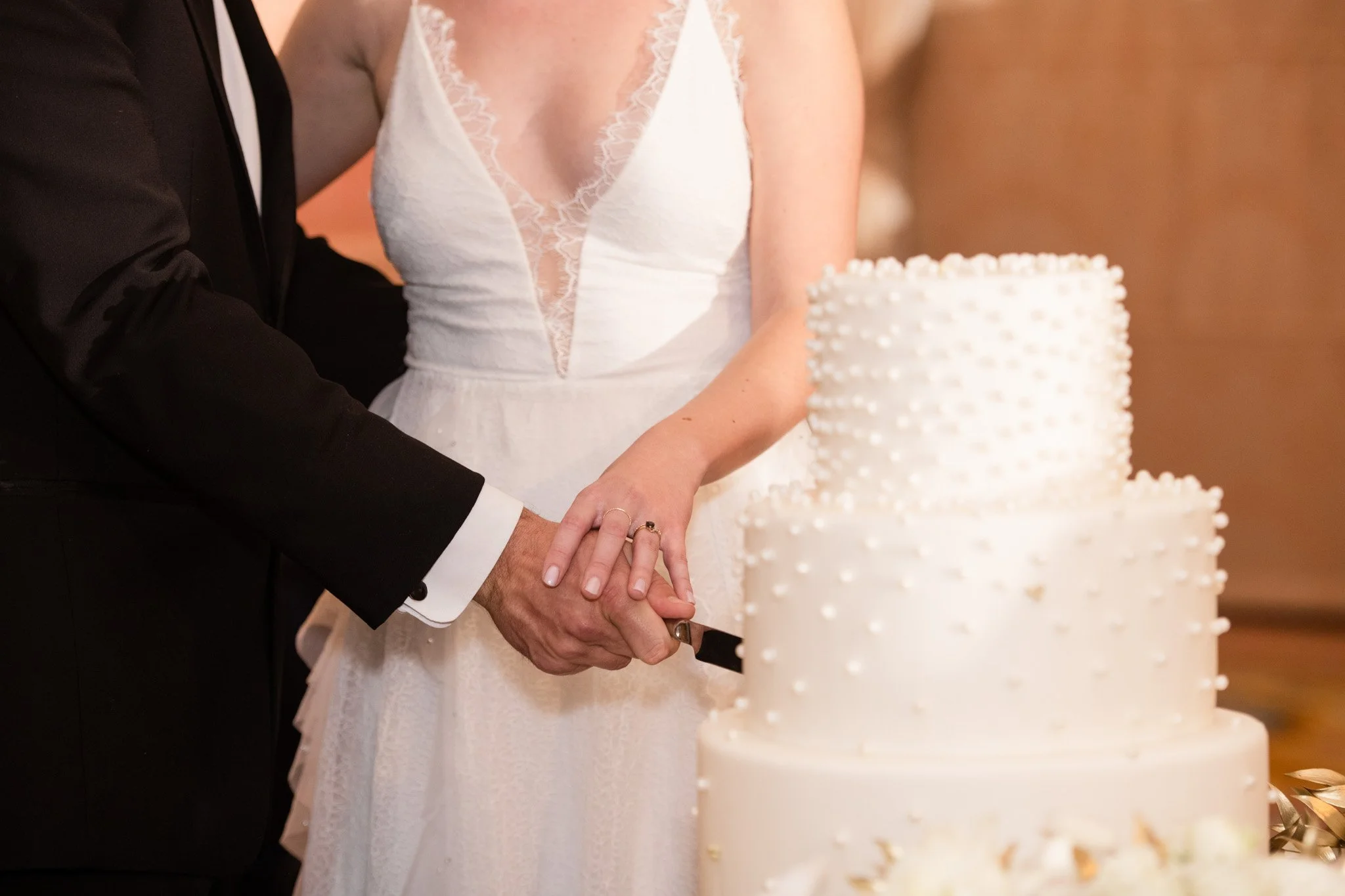 A newlywed couple cutting a white, multi-tiered wedding cake decorated with small white dots.