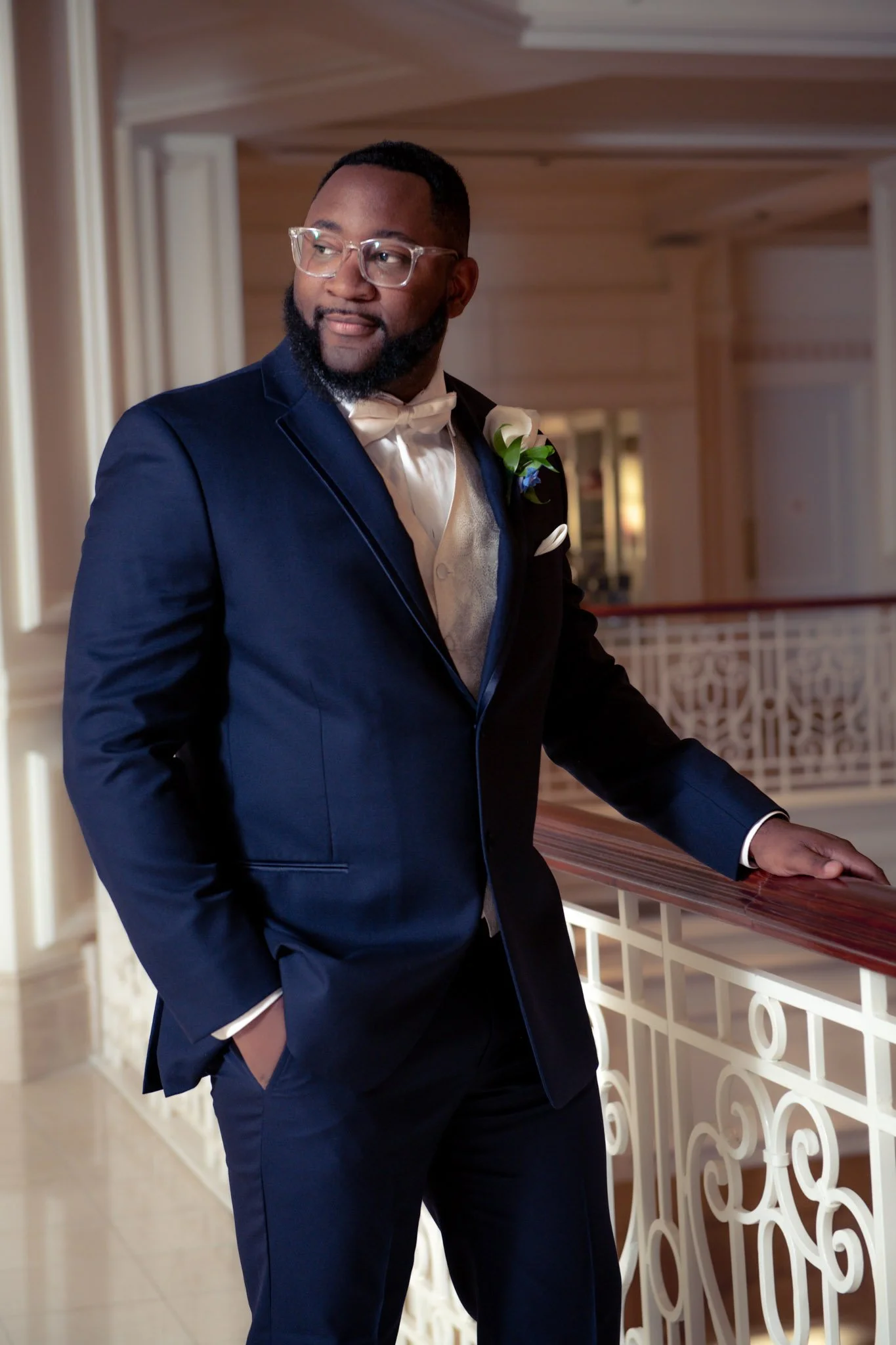 A man in a navy blue tuxedo with a white shirt, cream-colored vest, and bow tie, standing on a balcony at a formal event, looking to the side with one hand in his pocket, wearing glasses and a boutonnière on his lapel.