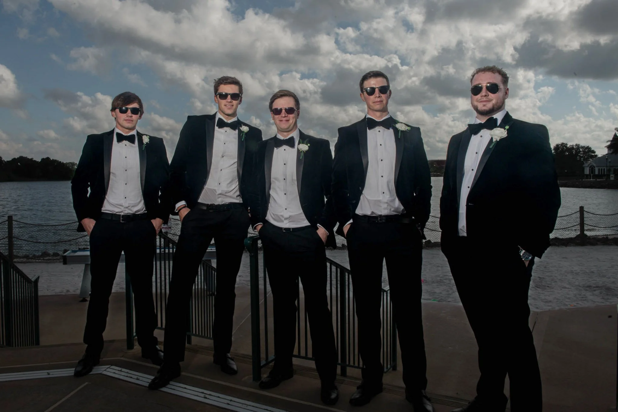 Six men in tuxedos and sunglasses standing outdoors near a body of water under a partly cloudy sky.