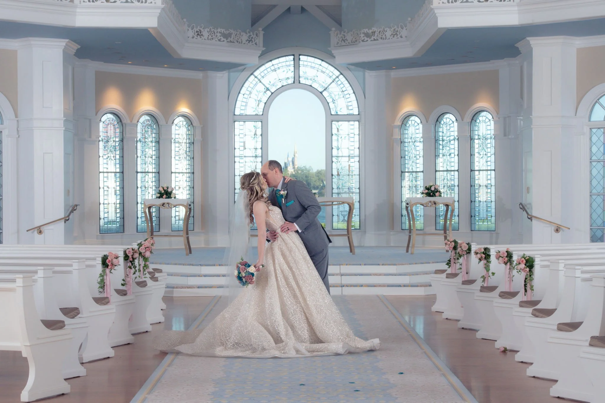 A bride and groom sharing a kiss in a beautifully decorated Disney Wedding Pavilion with large windows, floral arrangements, and elegant chairs.