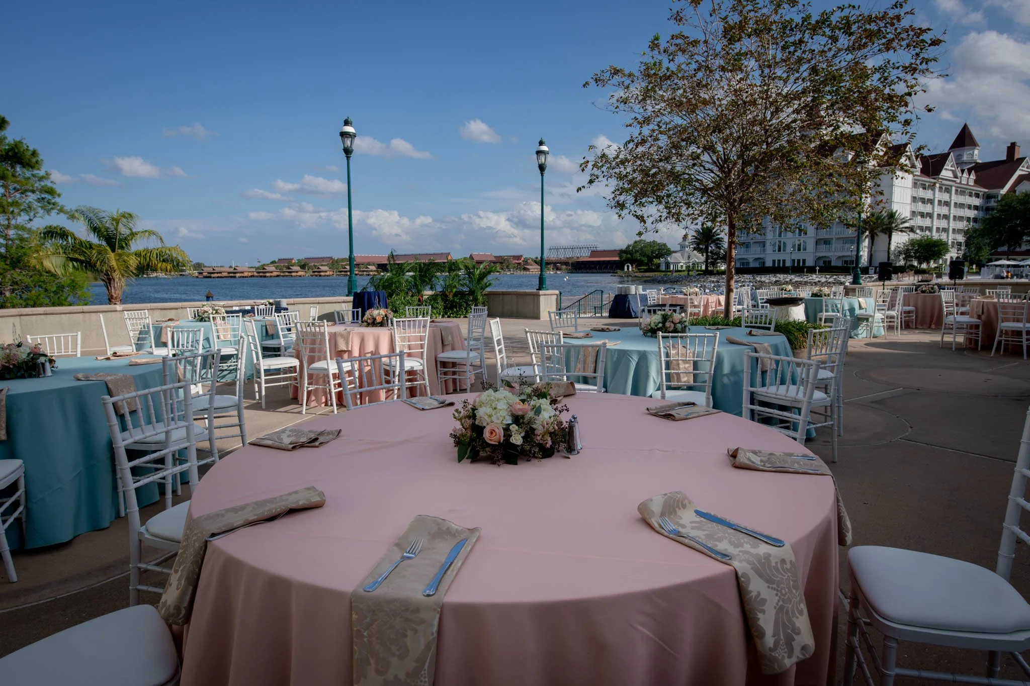 Outdoor event setup with round tables covered in pastel pink and blue tablecloths, decorated with floral centerpieces, surrounded by white chairs, overlooking a body of water under a partly cloudy sky at The Grand Floridian.