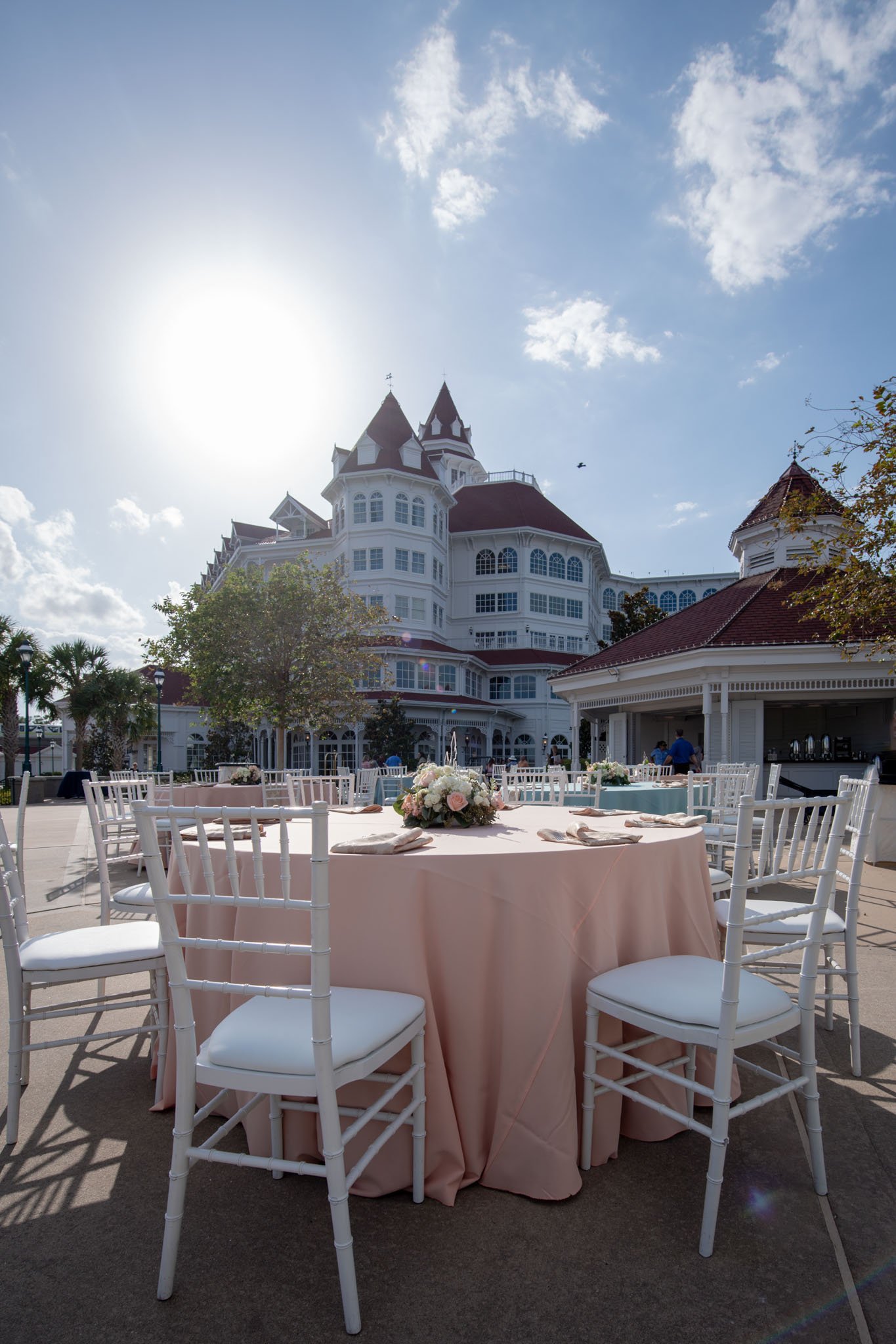 Outdoor setting with a round table covered in a pink tablecloth, decorated with a floral centerpiece, surrounded by white chairs, in front of The Grand Floridian with multiple turrets, windows, and balconies under a sunny sky 
